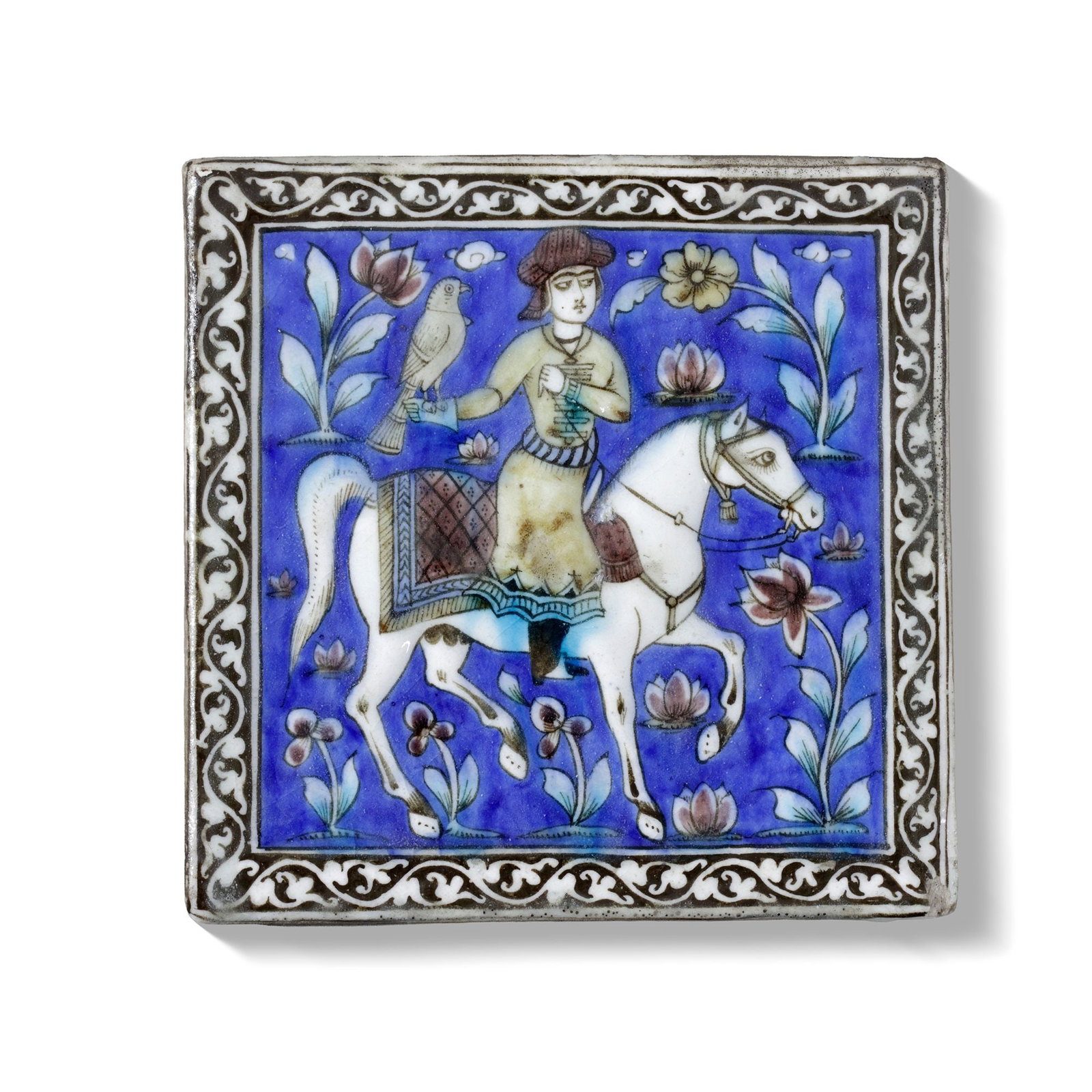 A POLYCHROME MOULDED POTTERY TILE, PERSIA QAJAR 19TH CENTURY: A Qajar ceramic tile depicts a poised horseman carrying a falcon, rendered in delicate underglaze tones of cobalt blue, turquoise, and green. The rider, shown in profile on a white horse, is set withi
