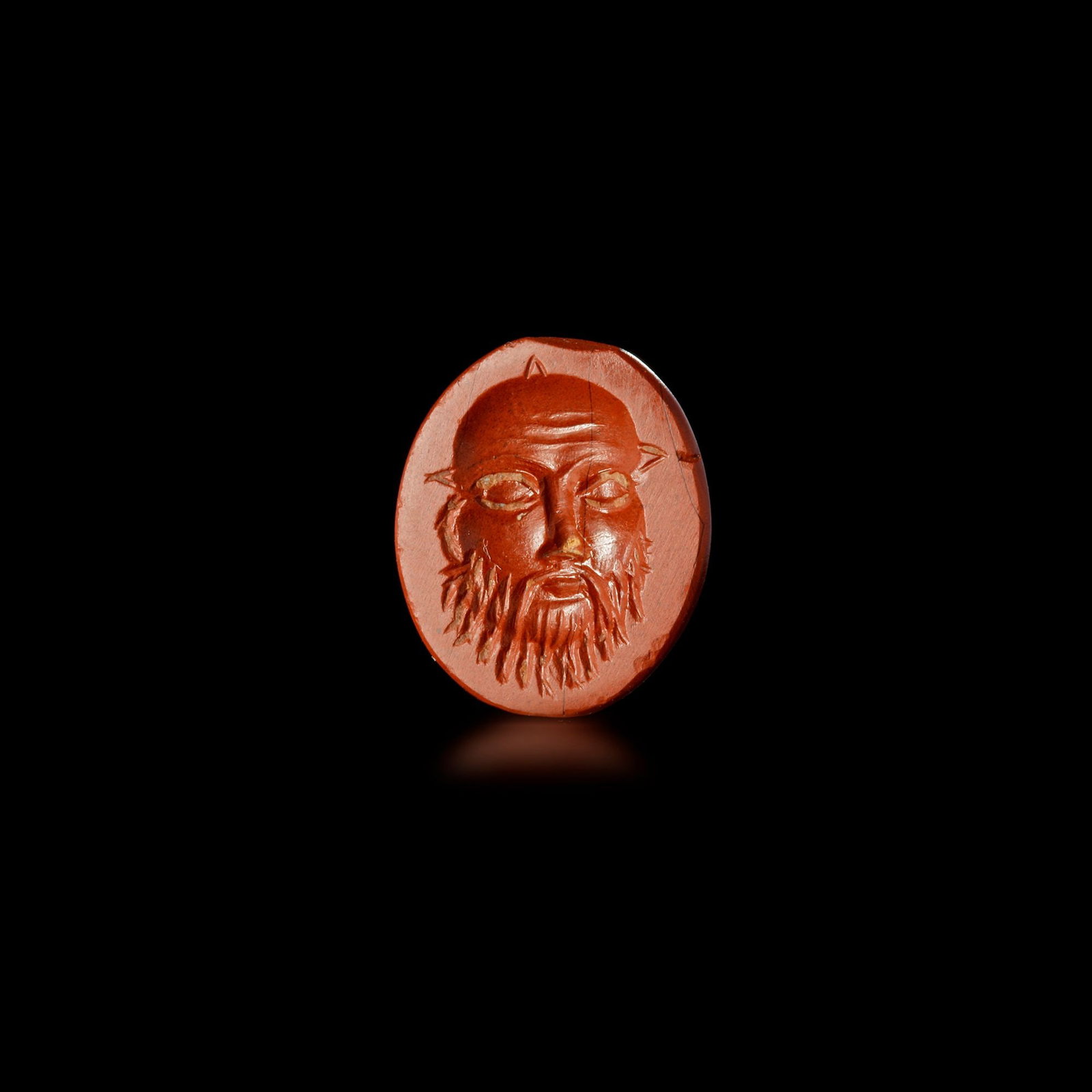 RED JASPER INTAGLIO OF SILENUSâ€™ FACE ROMAN, 1STâ€“2ND CENTURY A.D.: This striking intaglio, carved from deep red jasper, presents the bearded face of Silenus, a companion and tutor to the god Dionysus in Greek and Roman mythology. Rendered with expressive detail, the