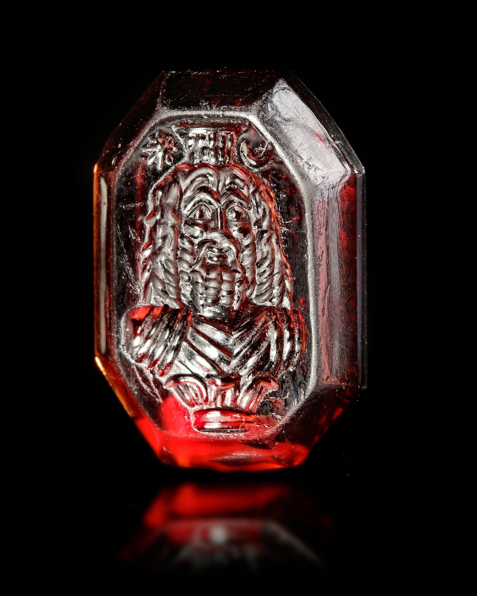 A ROMAN GARNET INTAGLIO OF SERAPIS, 1ST-2ND CENTURY AD: An octagonal intaglio in garnet showing a facing bust of Serapis flanked by the moon and a star. Garnet, Roman,1/2nd.c.A.D. 19 by 14 mm. Weight: 2.1 gr.PROVENANCE From a European collection since the