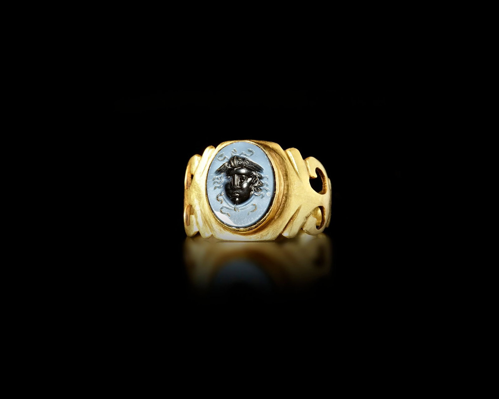 A ROMAN GOLD OPENWORK RING WITH NICOLO INTAGLIO, 2ND-3ND CENTURY AD: Roman gold ring featuring an elegant openwork shoulder design and set with a finely engraved nicolo intaglio depicting Medusa. The contrast between the pale and dark layers of the nicolo stone highlig