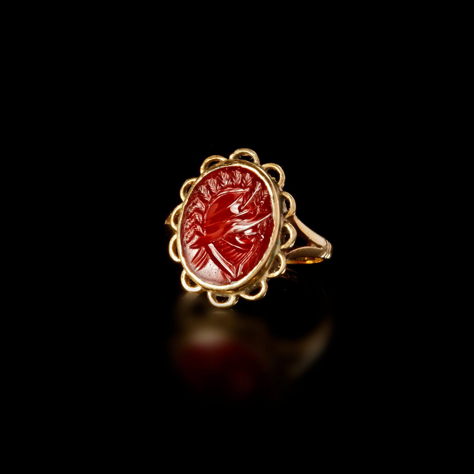 A ROMAN AGATE INTAGLIO RING DEPICTING A MILITARY HELMET â€“ 1ST CENTURY A.D.: This elegant Roman ring,features a finely carved agate intaglio set within an ornate gold bezel of scalloped loops. The vivid red stone is engraved with a striking depiction of a Roman military helmet