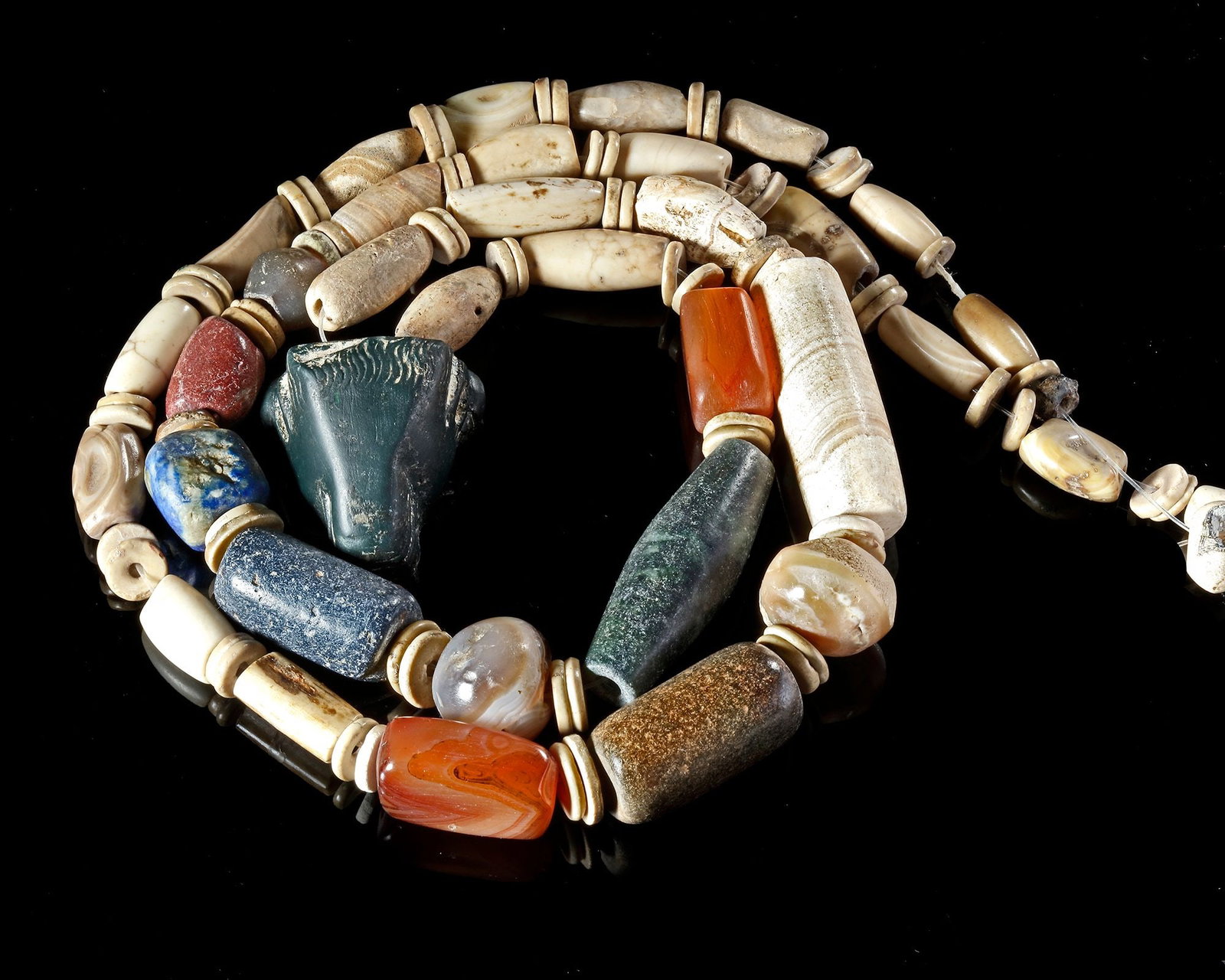 A NECKLACE OF BEADS WITH BULL HEAD PENDANT, 2ND-3RD MILLENNIUM BC: A necklace composed of stone and shell beads of various periods, mainly pre-dynastic Akkad and other Bronze Age cultures. The pendant of a bull's head is rendered in a very fine dark green stone and i