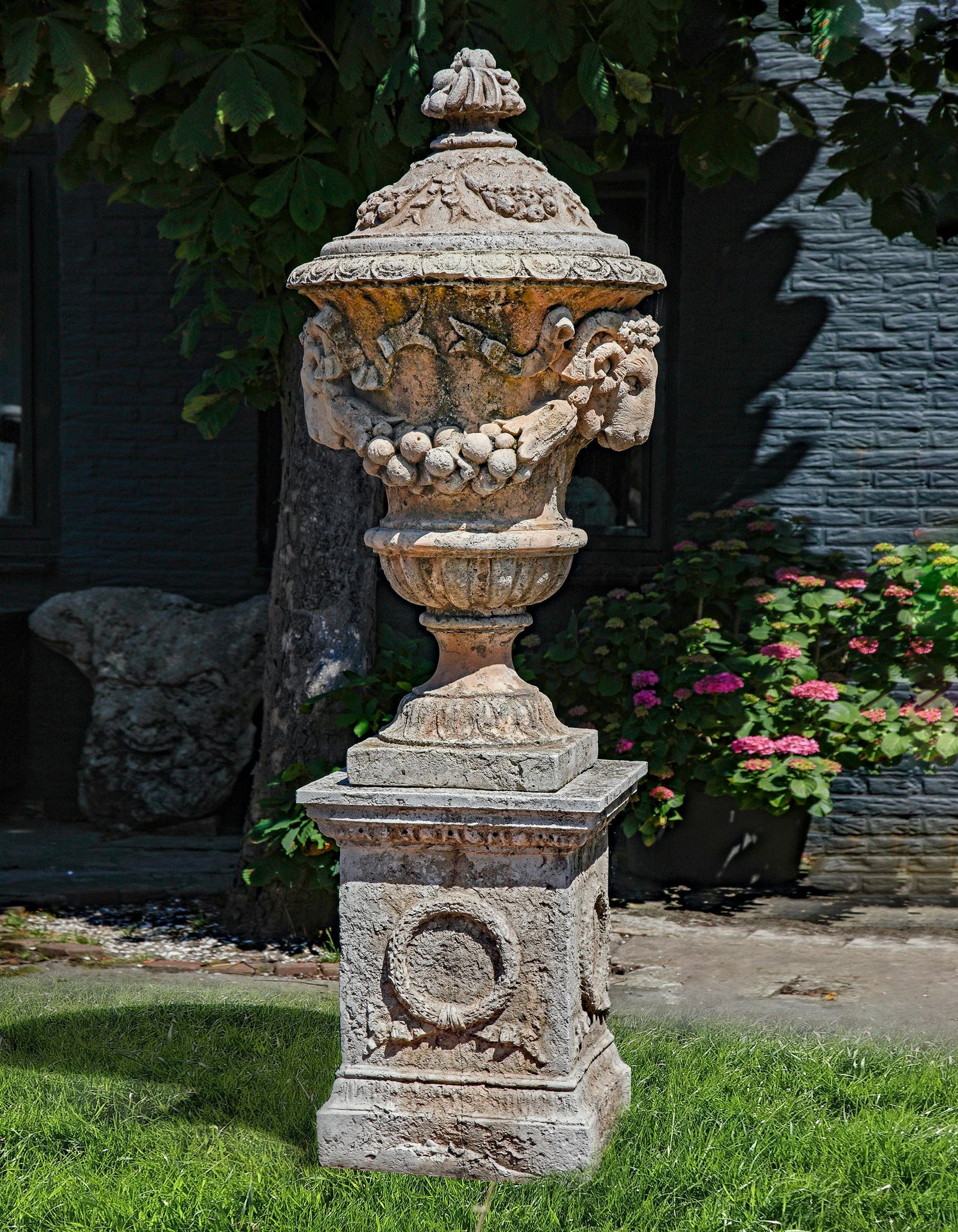 A COMPOSITION STONE GARDEN URN IN 18TH CENTURY TASTE, SECOND HALF 20TH CENTURY: With applied twin ramâ€™s heads and swag decoration, height 143 cm. Atop an associated square section pedestal base. Height 73.5 cm. Width 51 cm. Depth at the base 53 cm. Height overall 216.5 
