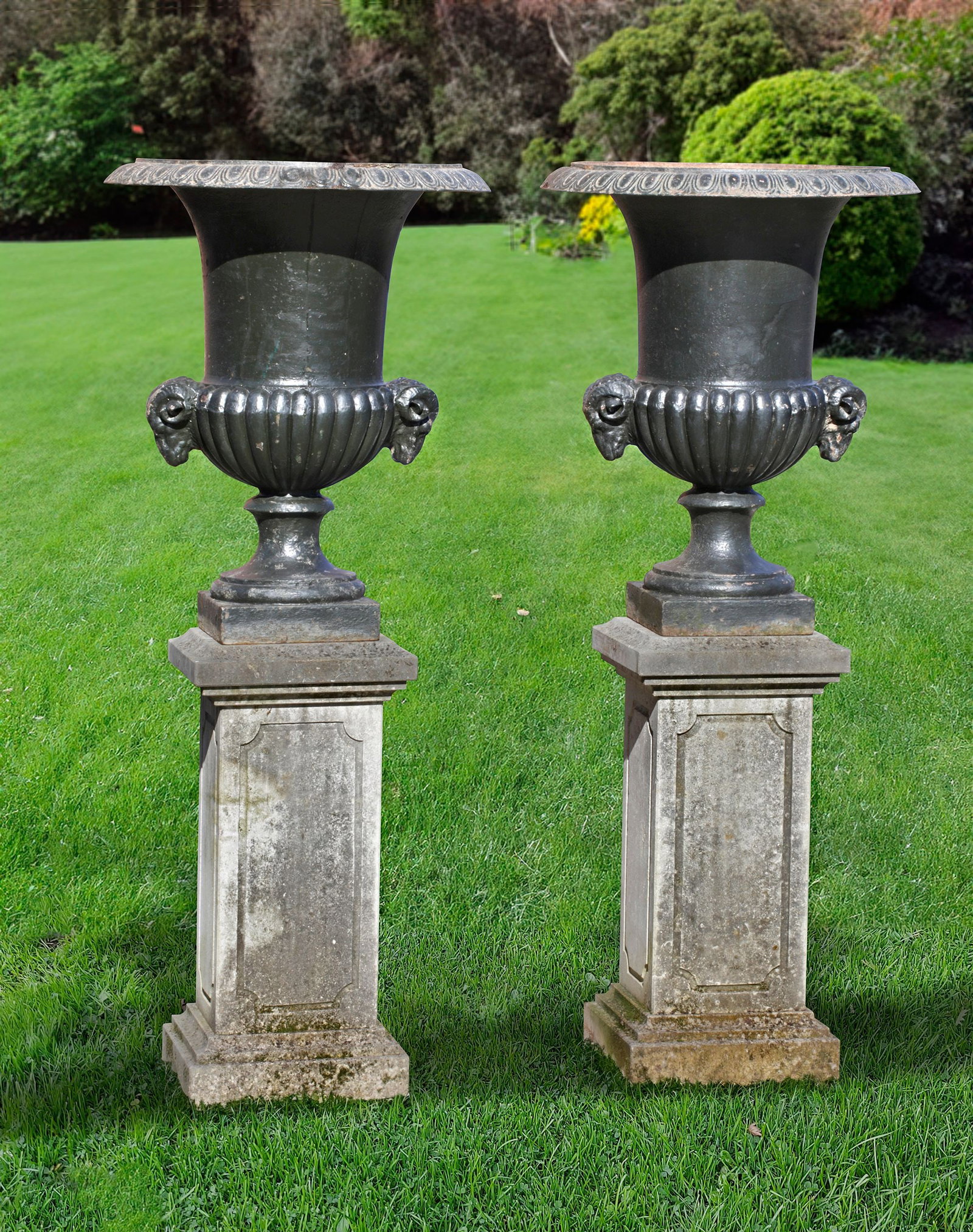 A PAIR OF CAST IRON CAMPANA URNS, 19TH CENTURY: With rams heads on the side, on later carved limestone pedestals. Height overall 144 cm. Diameter 60 cm. Base 71 by 34 by 34 cm.