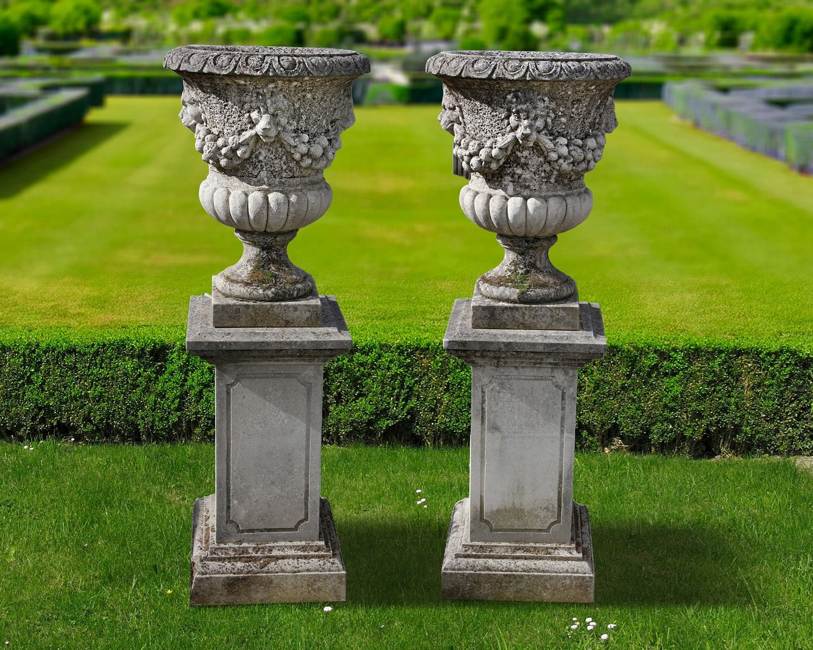 A PAIR OF LIMESTONE GARDEN URNS, LATE 19TH OR EARLY 20TH CENTURY: Each with an everted egg-and-dart rim above the body with stylised lion masks issuing swags and lobed lower portions. Height each 72 cm. Diameter 55 cm. Atop square section pedestals, height 80 cm. an