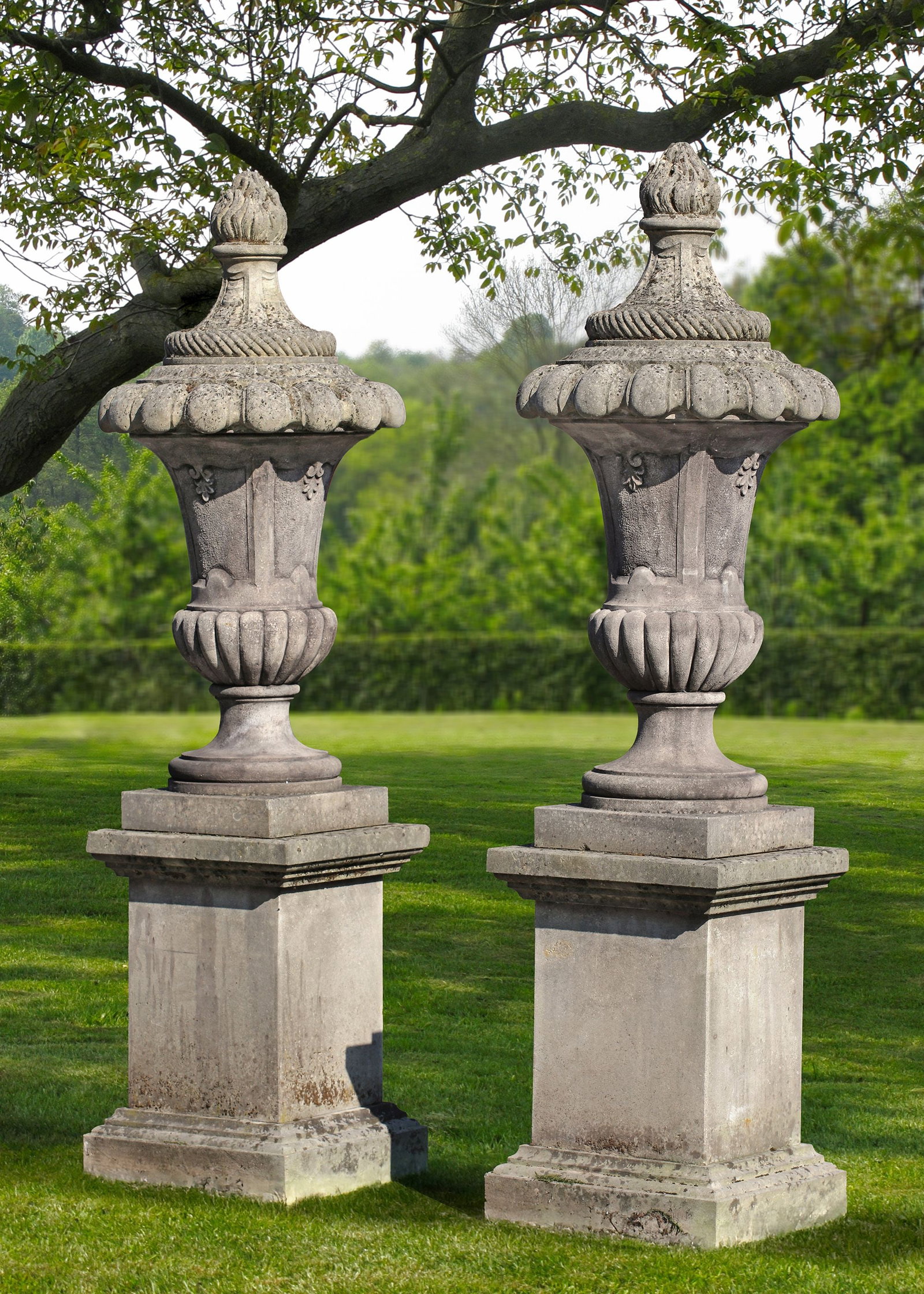 A PAIR SCULPTED LIMESTONE GARDEN VASES ON TALL PEDESTALS, 20TH CENTURY: The central part of the vase is slimmer and decorated with vertical lines and subtle relief ornaments, the top rim has a decorative band with a twisted motif, and above it a wreath of rounded shapes. 
