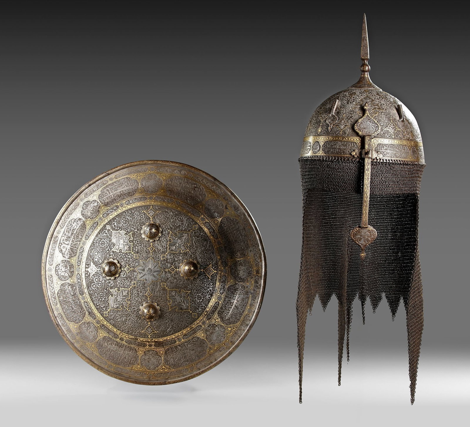 A GOLD-DAMASCENED STEEL HELMET AND SHIELD, QAJAR, PERSIA, EARLY 19TH CENTURY: Both engraved and decorated in gold overlay with inscription-filled cartouches, with openwork decoration interspersed by panels with foliate designs, the helmet of domed form surmounted by a spike fin