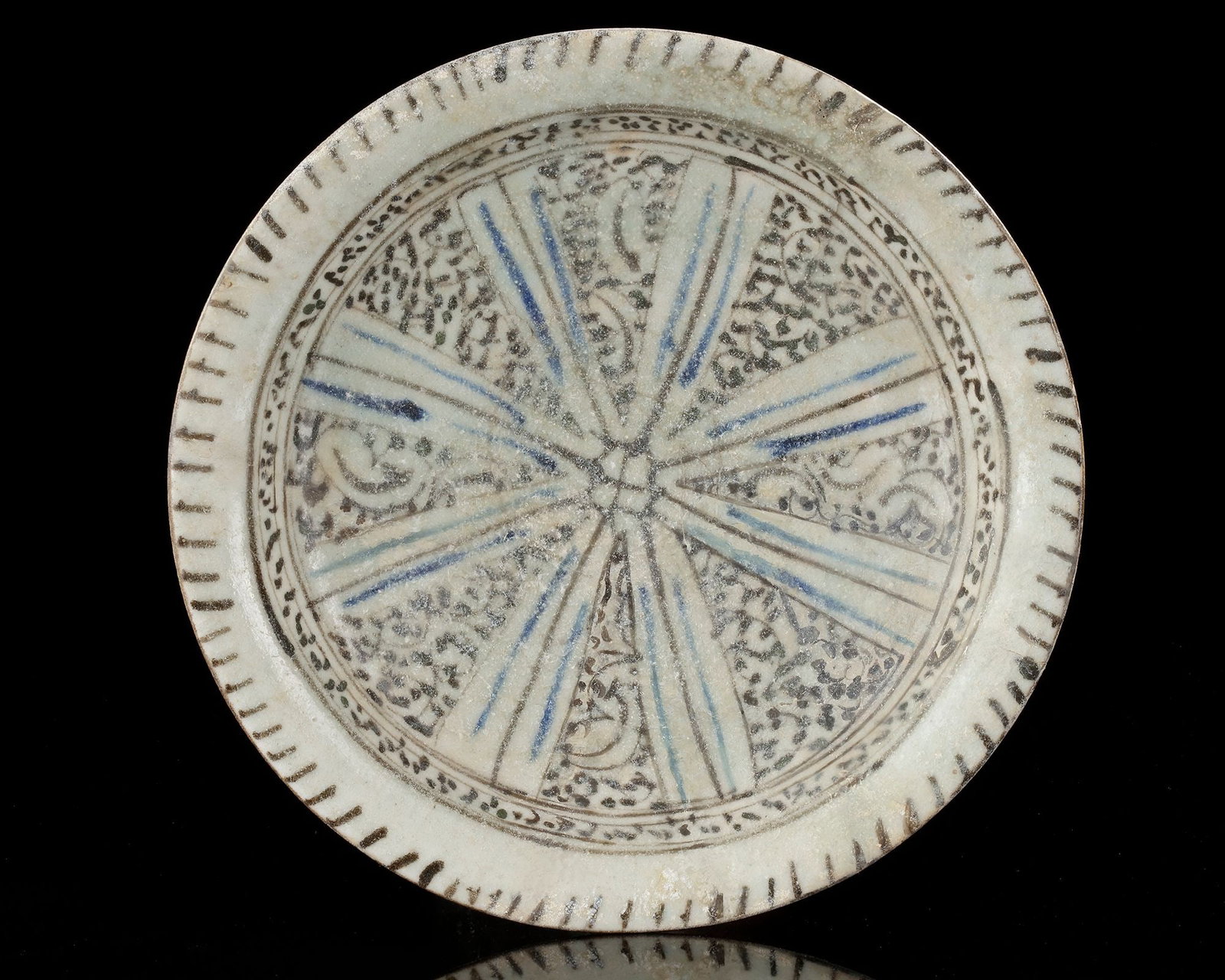 A KASHAN BLUE, BLACK AND WHITE POTTERY BOWL, PERSIA, EARLY 13TH CENTURY: A Pottery broad, shallow circular bowl set on a tall cylindrical foot. Inside, the decoration is arranged in a radial composition divided into triangular segments that spread outward from the center. 