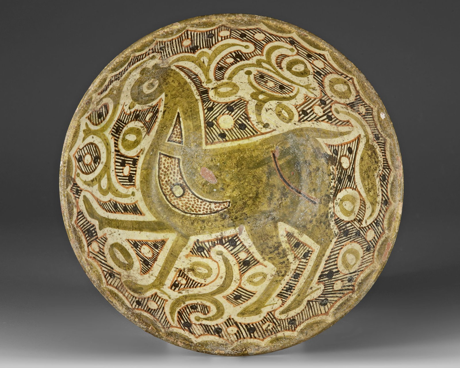 A NISHAPUR SLIP-PAINTED POTTERY BOWL, PERSIA 10TH-11TH CENTURY: A Nishapur slip-painted pottery bowl of shallow, flaring form on a low foot, the buff slip ground decorated in olive green, brown, and black pigments. The interior centers on a stylized quadruped rend