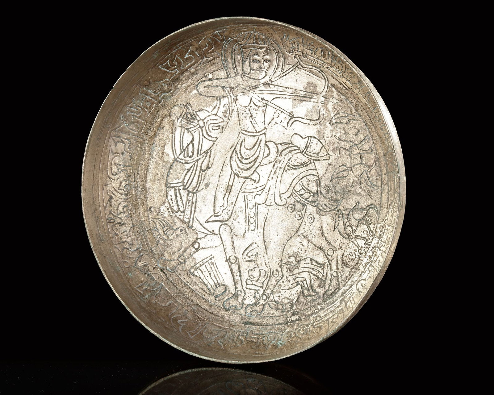 A GHAZNAVID BRASS BOWL WITH CAMEL, EASTERN PERSIA, 12TH CENTURY: The interior engraved with a central figural scene depicting a mounted rider on a camel, surrounded by animals and attendants. the composition is enclosed within a circular band of kufic inscription. 