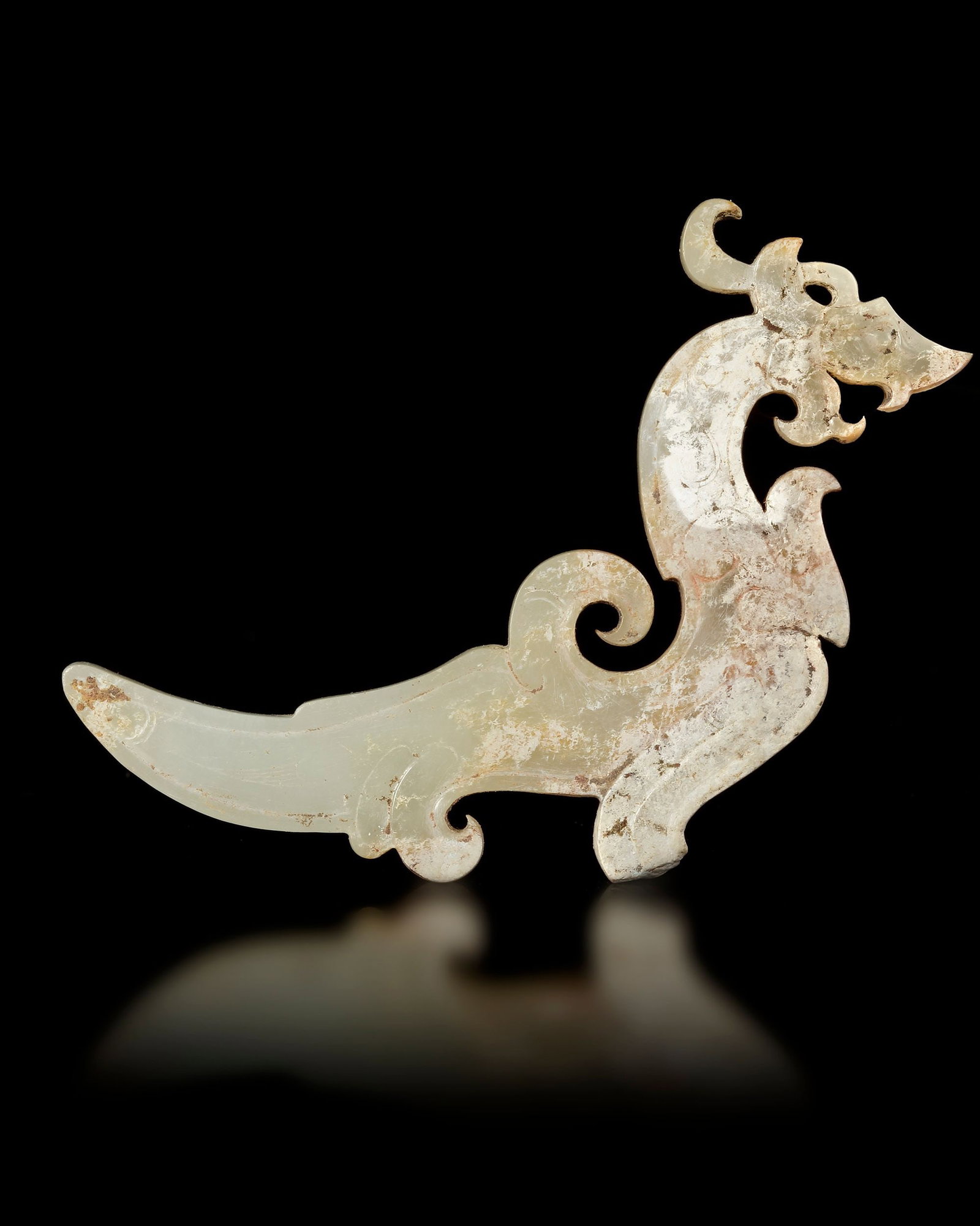 A JADE PENDANT, ZHOU DYNASTY (CIRCA 1046â€“256 BC): A finely carved jade pendant in the form of a stylized mythical creature. The piece features elegant flowing curves and openwork details typical of ancient Chinese jade ornaments. Subtle natural surfa