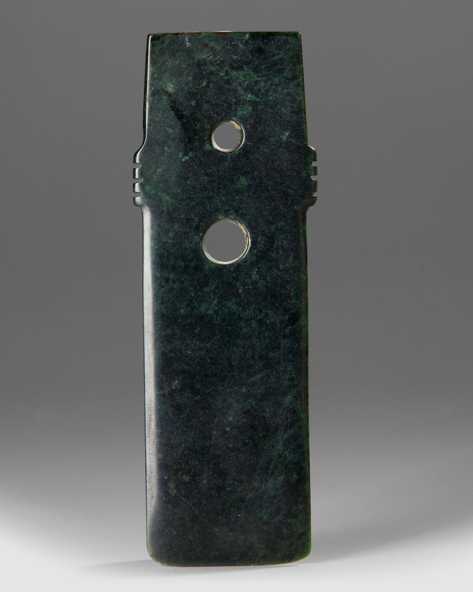 A GREEN JADE GUI TABLET, CHINA, SHANG DYNASTY (c. 1600â€“1046 BC): Ceremonial gui tablet carved from dark green jade, . The elongated rectangular form features two drilled circular perforations and small notched projections on the sides near the upper section, charac