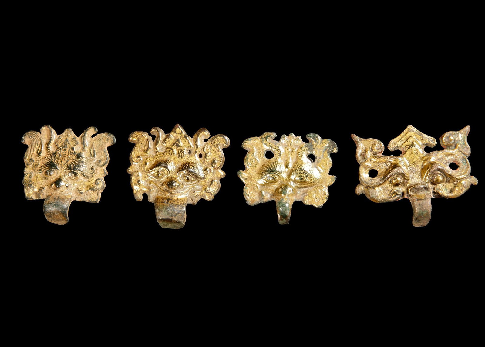 A SET FOUR OF GILT-BRONZE 'TAOTIE MASK' HANDLES, CHINA: Each crisply cast with two short horns flanking a pointed cranium, large bulbous eyes and a loop from the mouth, the front surface with extensive gilding remaining, stand. Each. 6 by 5 cm. and 5 by 4 