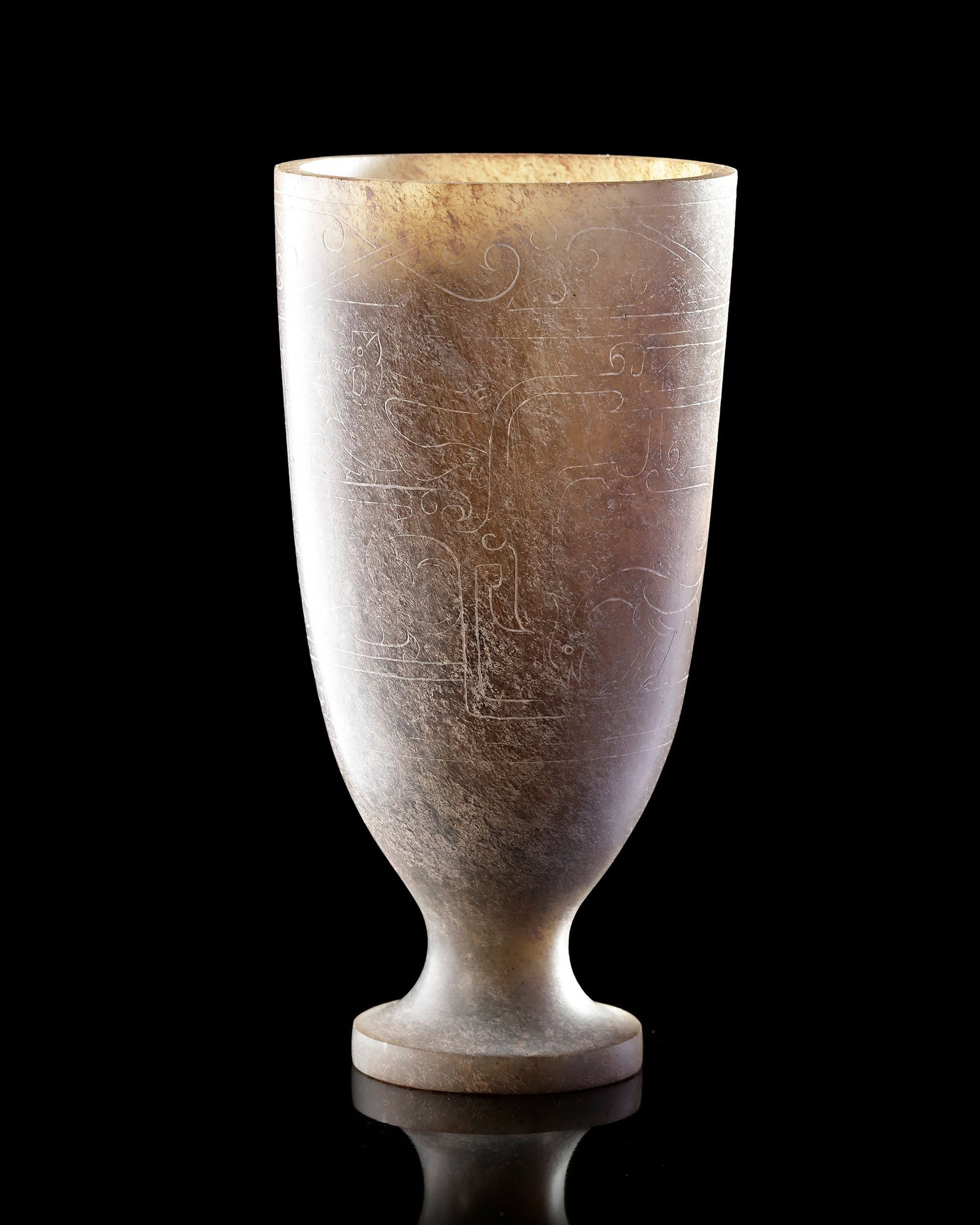 A CHINESE JADE BEAKER, HAN DYNASTY (206 BC â€“ 220 AD): A finely carved jade beaker raised on a small foot .The vessel has a tall, elegant form with a slightly flared rim and a tapering body. Delicate incised decoration with stylized scrolls and archaic mo