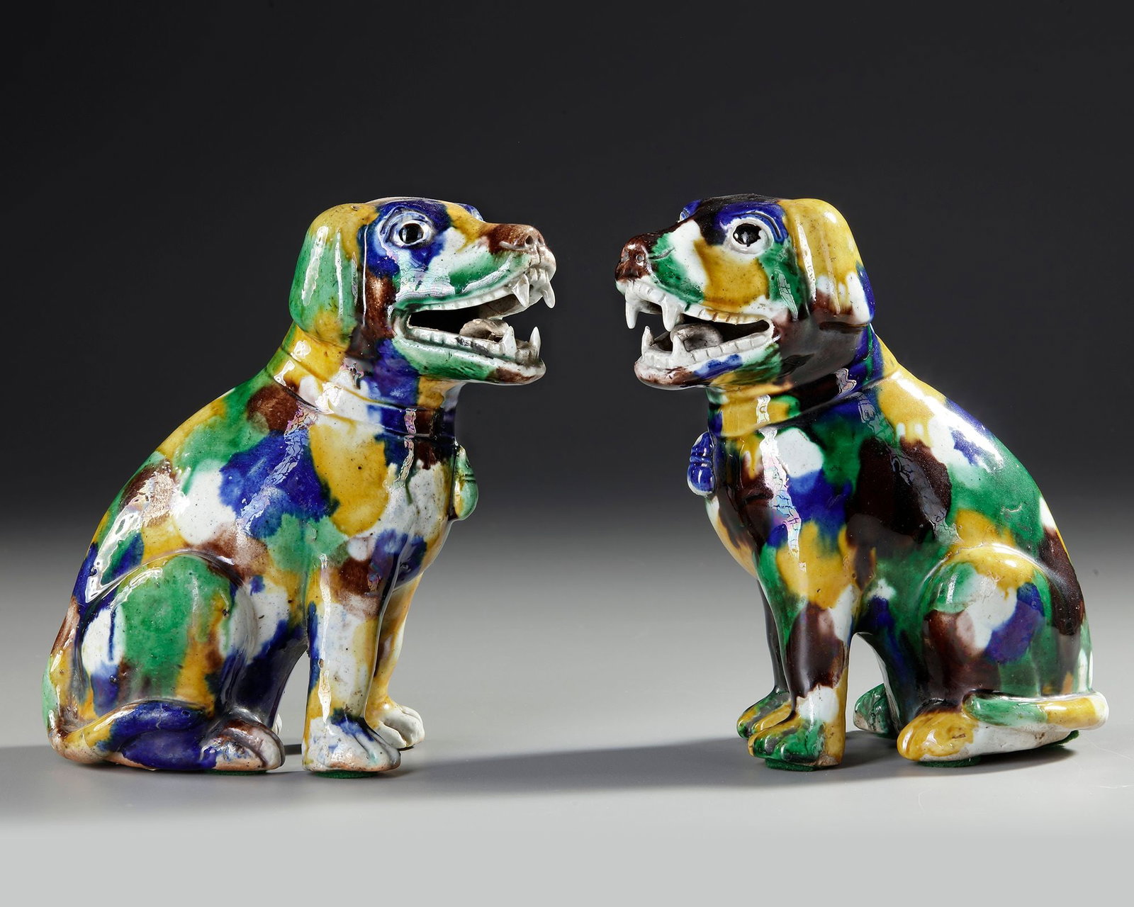 A PAIR OF CHINESE SANCAI GLAZED DOGS, 18TH CENTURY: A striking pair of Chinese sancai-glazed pottery dogs . Modelled seated with alert expressions and open jaws, the figures are covered in vibrant splashed glazes of green, yellow, blue, brown, and whit