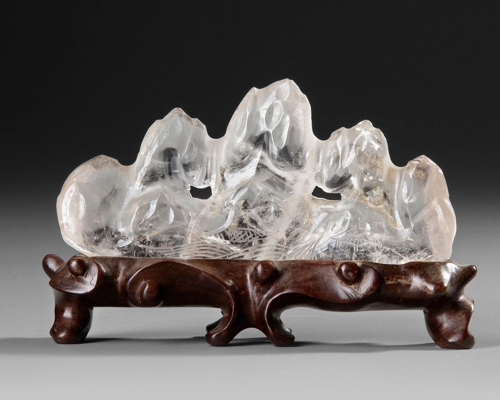 A CHINESE ROCK CRYSTAL BRUSH REST, 19TH CENTURY: An elegant Chinese rock crystal brush, finely carved in the form of a mountain landscape with soaring peaks and flowing contours. The translucent crystal is beautifully displayed on a carved hardwood 