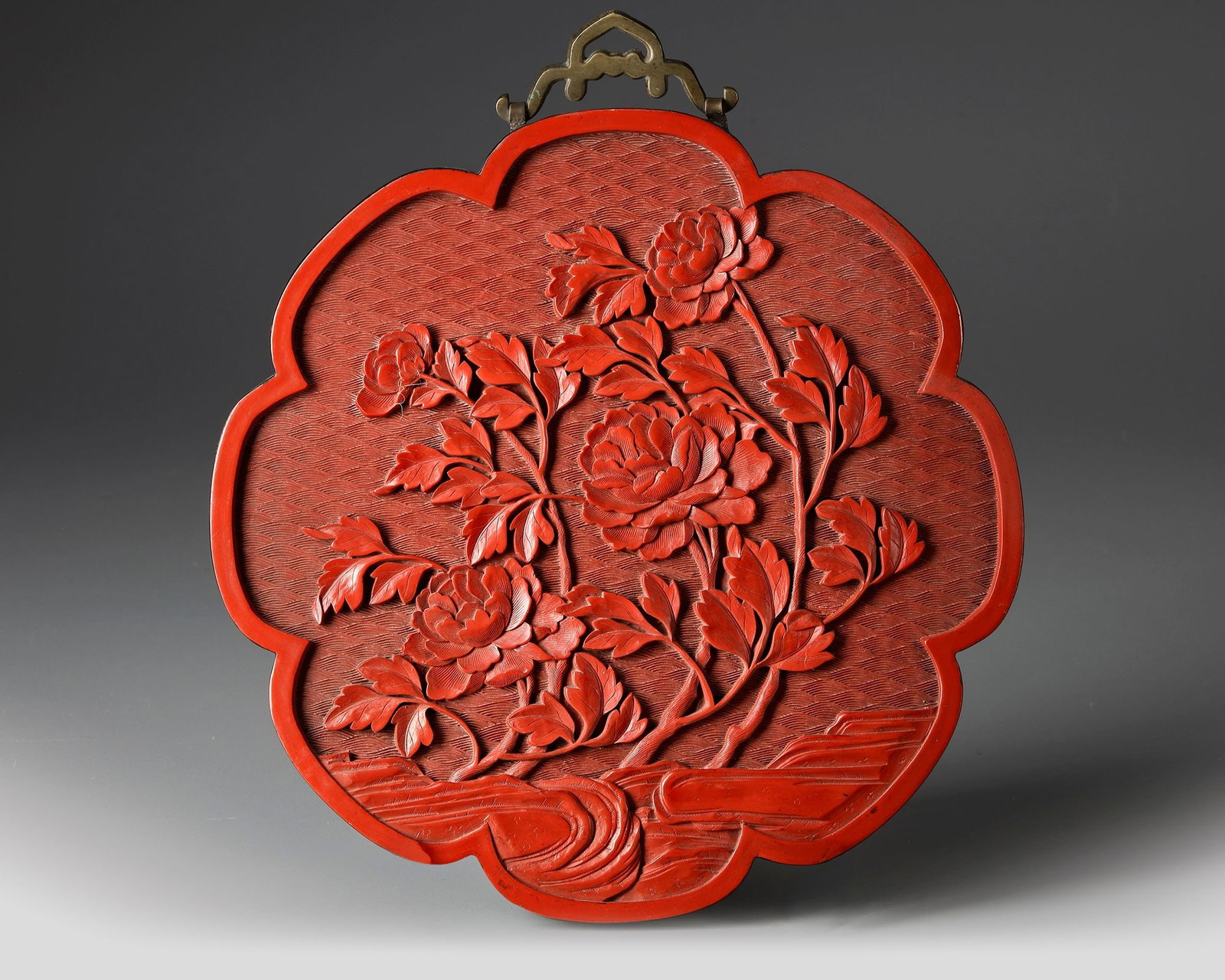 A CHINESE RED LACQUER PANEL 19TH CENTURY: A finely carved Chinese red lacquer panel of lobed floral form, decorated in high relief with blooming peony branches against a textured diaper-pattern ground. Diameter 25 cm. Provenance: former colle