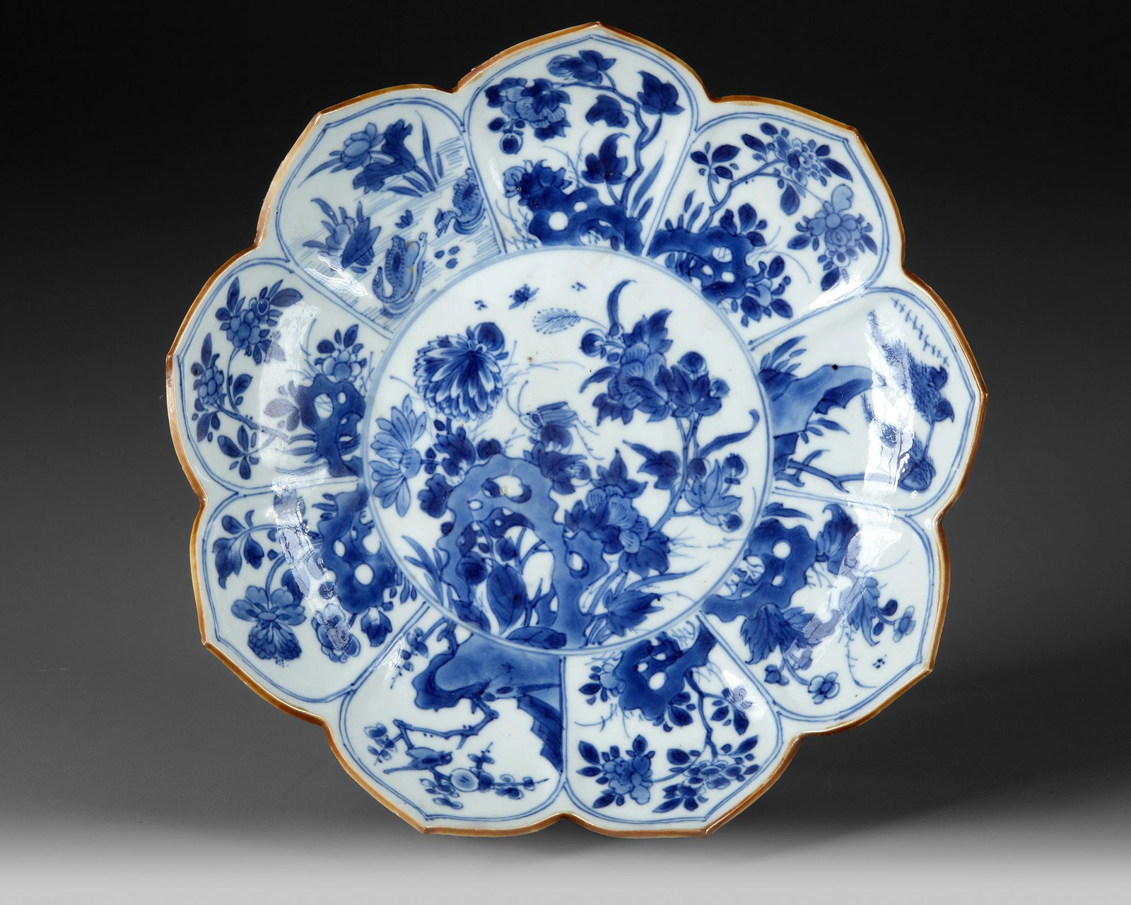 A CHINESE BLUE AND WHITE 'LOTUS' DISH, KANGXI PERIOD (1662-1722): With a rim divided into lobes and painted with birds in flowers, the central panel depicting a grasshopper among rocks and blossoms. Diameter: 22.5 cm. Restoration to one of the lotus.