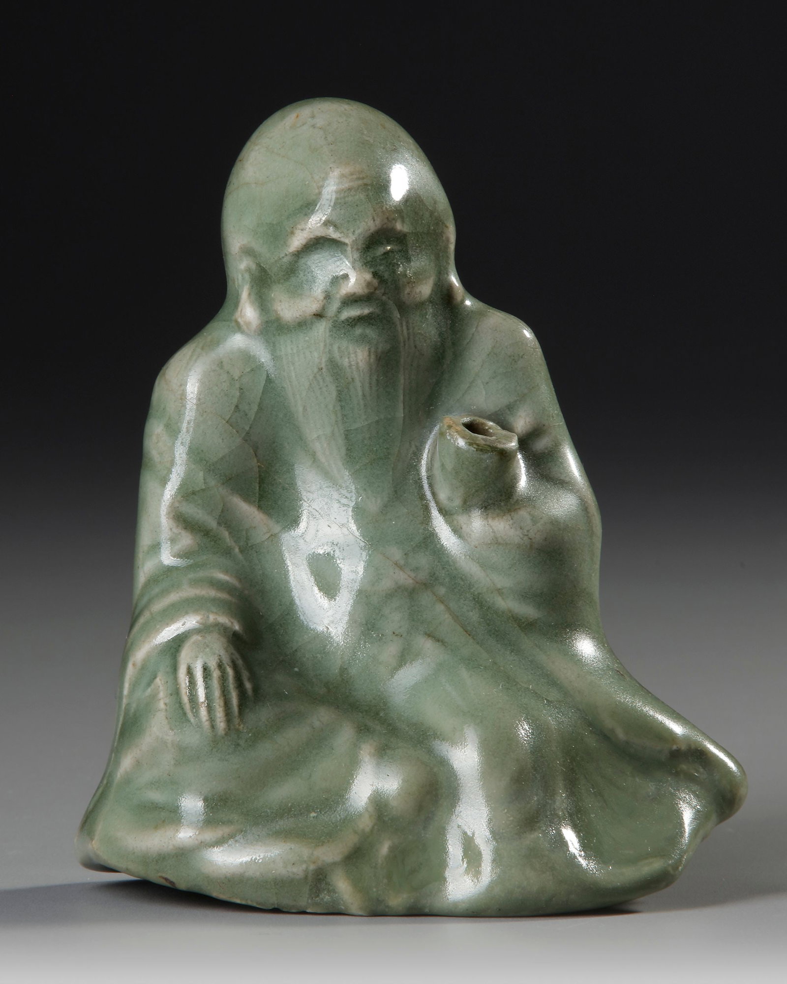A CHINESE CELADON FIGURE CENSER, MING DYNASTY, 15TH CENTURY: Chinese celadon-glazed figure of a seated sage, modeled as an incense burner. The elderly figure is depicted in flowing robes with an expressive face and elongated beard, holding a small vessel that s