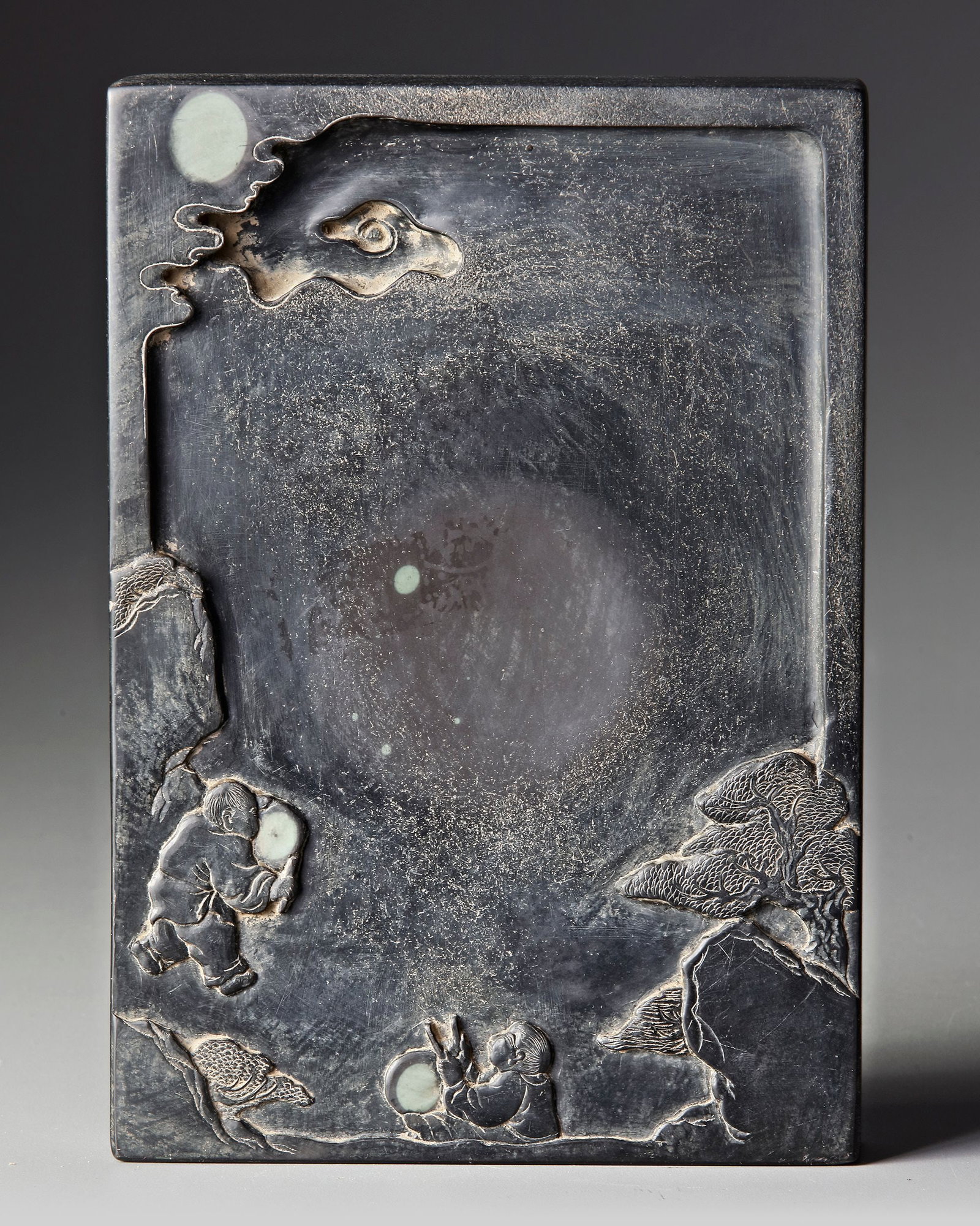 A CHINESE INKSTONE, QING DYNASTY (1644-1912): A finely carved Chinese inkstone , crafted from dark stone with a smooth grinding surface. The piece is decorated with a delicate relief scene featuring small figures in a landscape with rocks, trees,