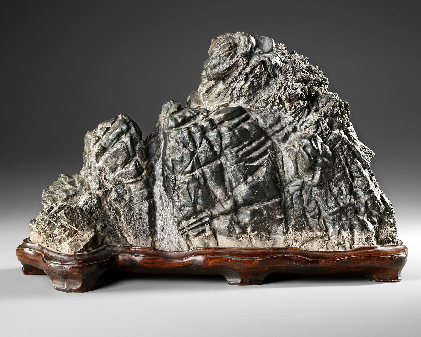 A LIU BO MOUNTAIN SCHOLARs ROCK, CHINA, QING DYNASTY (1644-1912): A striking Liu Bo mountain scholars rock carved from dark stone, displaying dramatic natural textures and rugged peaks reminiscent of a mountainous landscape. The sculptural form reflects the traditio