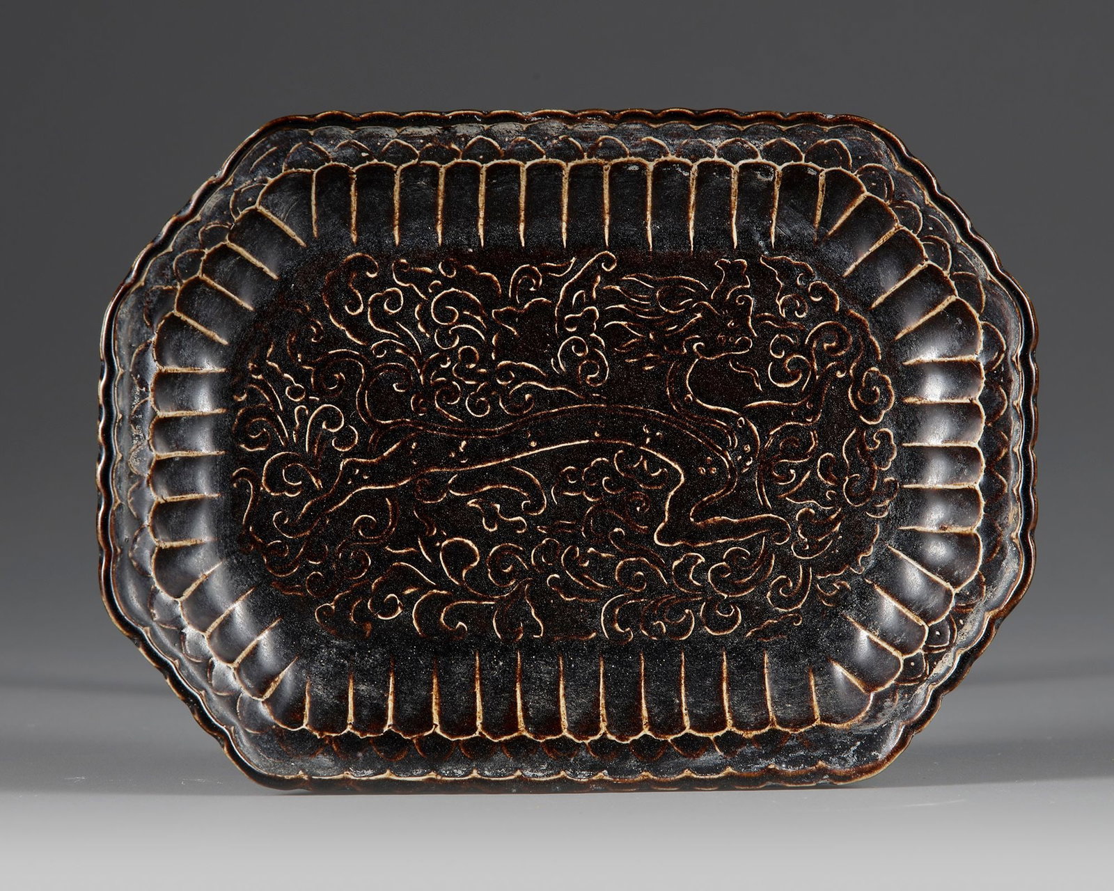 A CHINESE CIZHOU-TYPE BLACK-GLAZED INCISED TRAY, SONG DYNASTY (960-1279): Elegantly potted as a shallow rectangular tray with rounded corners and a lobed rim, the interior finely incised through a lustrous black glaze with a dynamic dragon amid swirling cloud scrolls. The d