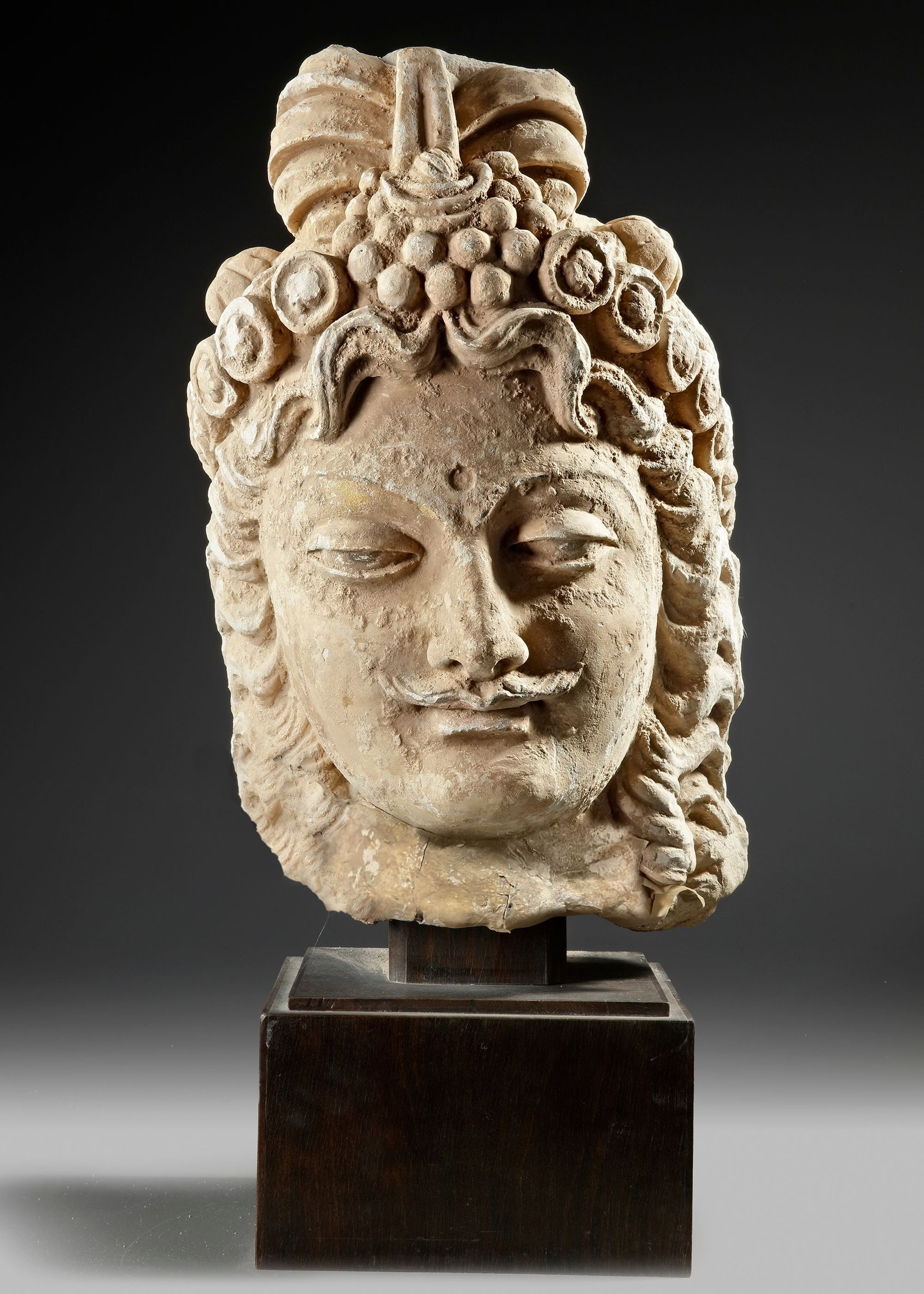 A GANDHARA HEAD, BUDDHIST SCULPTURE: A large Gandharan stucco head representing a serene Buddhist figure, mounted on a wooden base. The sculpture displays characteristic features of Gandhara art, including softly modeled facial features,