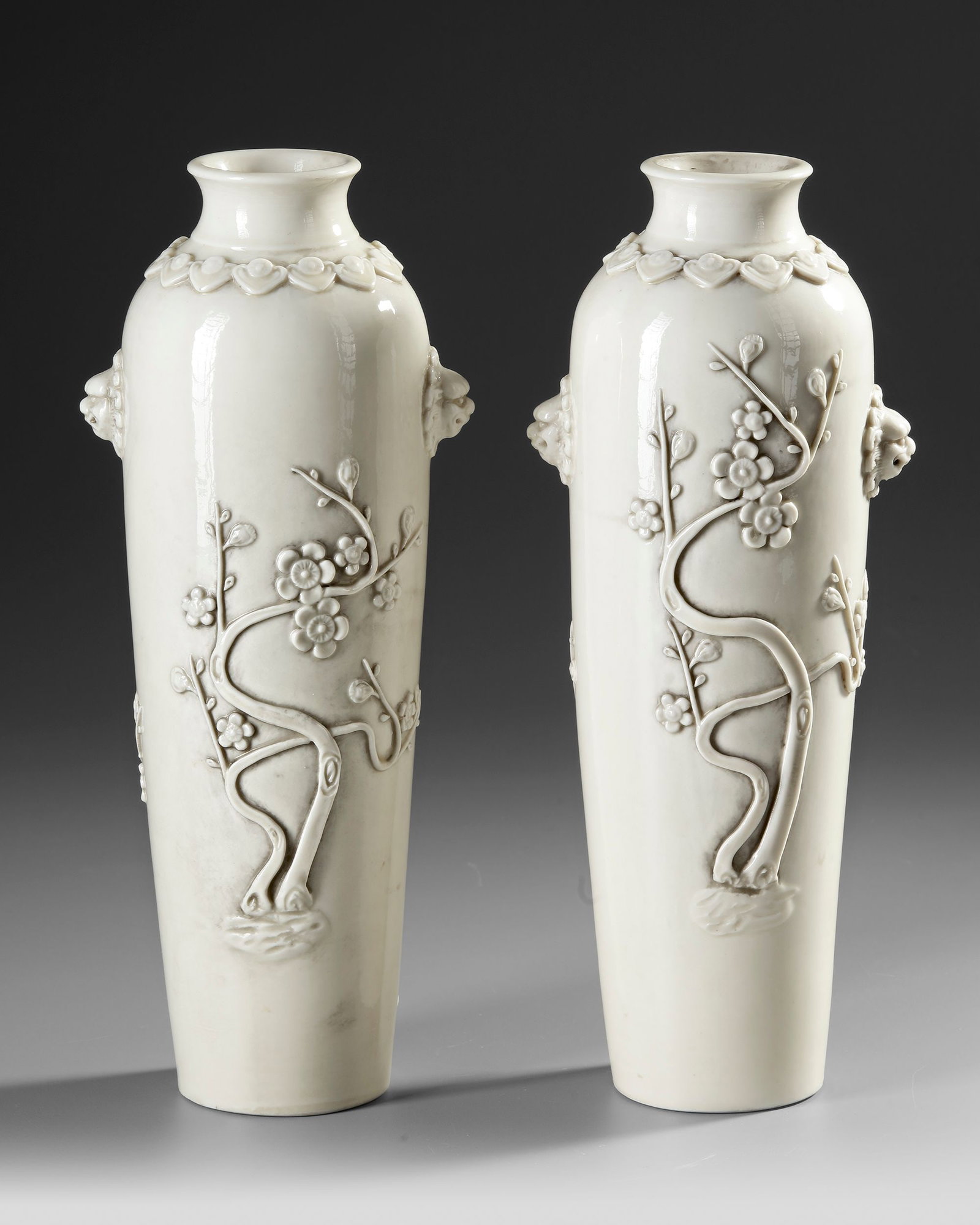 A PAIR OF CHINESE DEHUA BLANC DE CHINE ROULEAU VASES,QING DYNASTY (1644â€“1911): Each vase is of slender, tapered cylindrical form with rounded shoulders, applied with pierced lion-mask handles. The front is decorated with upright prunus branches, and a band of ruyi heads encircle