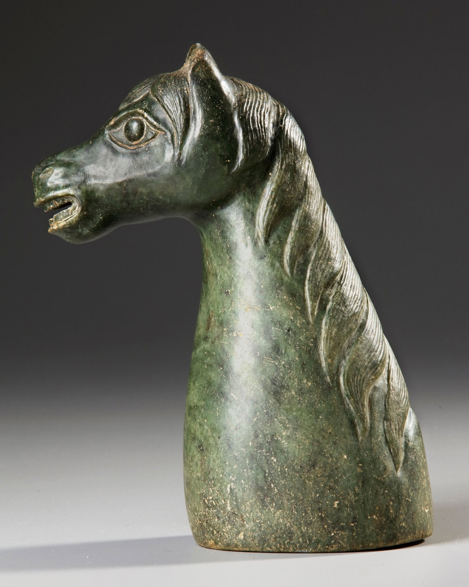 CHINESE JADE HORSE HEAD, 19TH CENTURY: A finely carved Chinese jade horse head . Sculpted from mottled green jade with natural inclusions, the piece displays expressive eyes, detailed mane work, and a powerful, elegant neck. height: 23 cm.