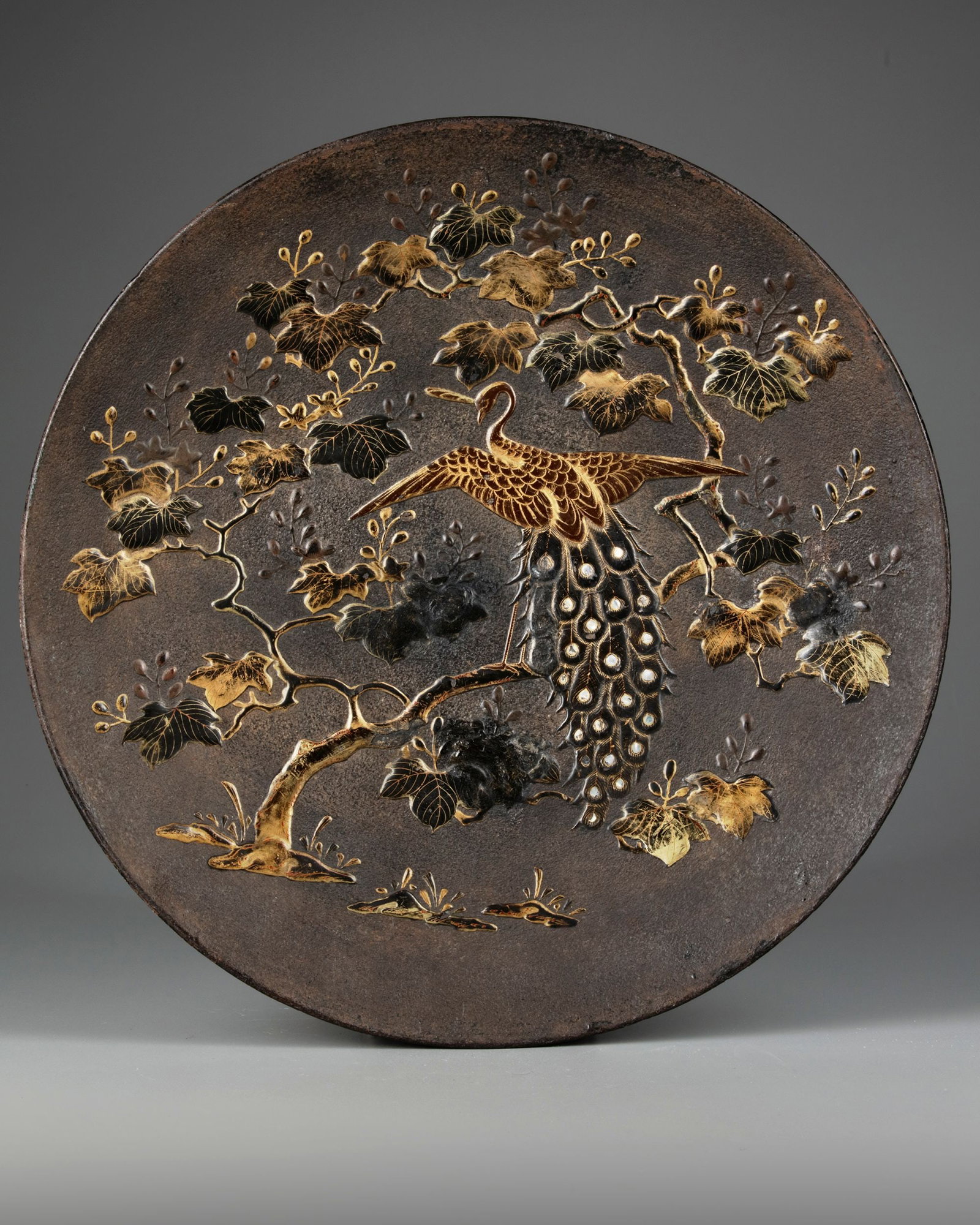 A JAPANESE METAL CHARGER, MEIJI PERIOD (1868-1912): A fine Japanese mixed metal charger featuring a majestic gilded peacock perched on a tree branch surrounded by intricately detailed gilded tree leaves. Diameter 39 cm.