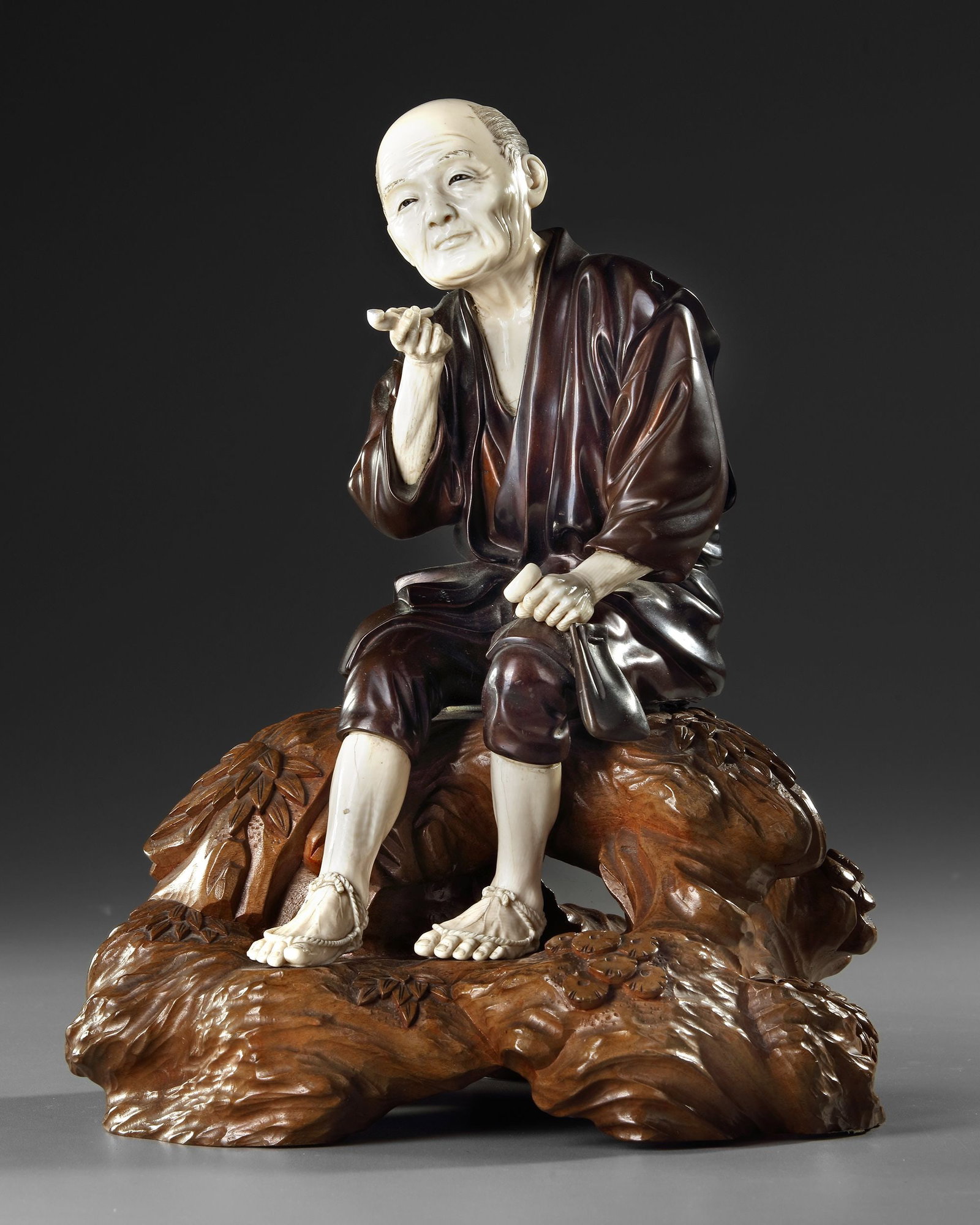 A LARGE JAPANESE BRONZE AND BONE OKIMONO OF A PIPE SMOKER, MEIJI PERIOD (1868-1912): Modelled seated on a wood stand, his right hand holding a pipe, his left a tobacco pouch. Height: 32 cm. with stand, 27 cm. without stand