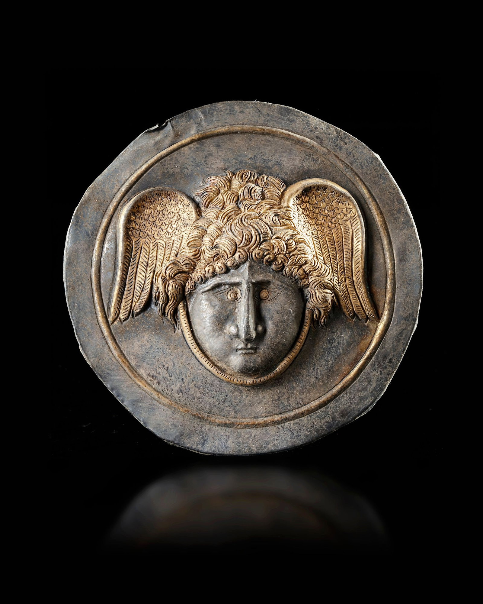 A ROMAN GILT SILVER PHALERA WITH WINGED HEAD OF MEDUSA, 1ST-2ND CENTURY AD: A Roman silver gilt phalera with a facing Gorgon head rendered in repouse, with a circular frame. A well known type, often used in decorations the cuirass used by Roman officers from the 1-3rd.c.A.D. 