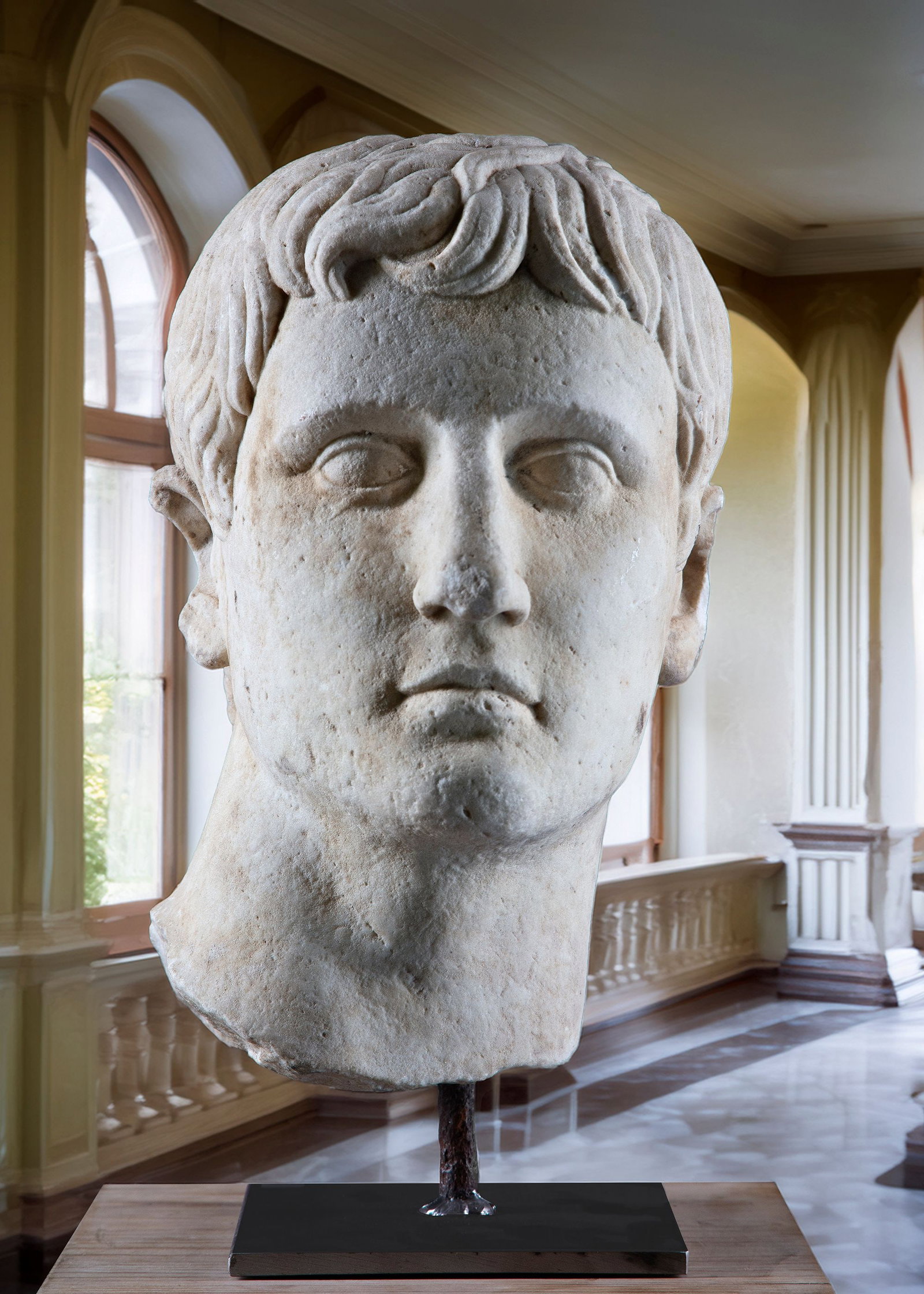 A GRAND TOUR SCULPTED WHITE MARBLE HEAD 19TH CENTURY, AFTER THE MANNER OF ROMAN EXAMPLES: Modelled looking ahead, with hair falling across the forehead, the rear socketed, now mounted on a modern wrought iron display stand. Height overall 48.5 cm. The base 19.2 by 20 cm.