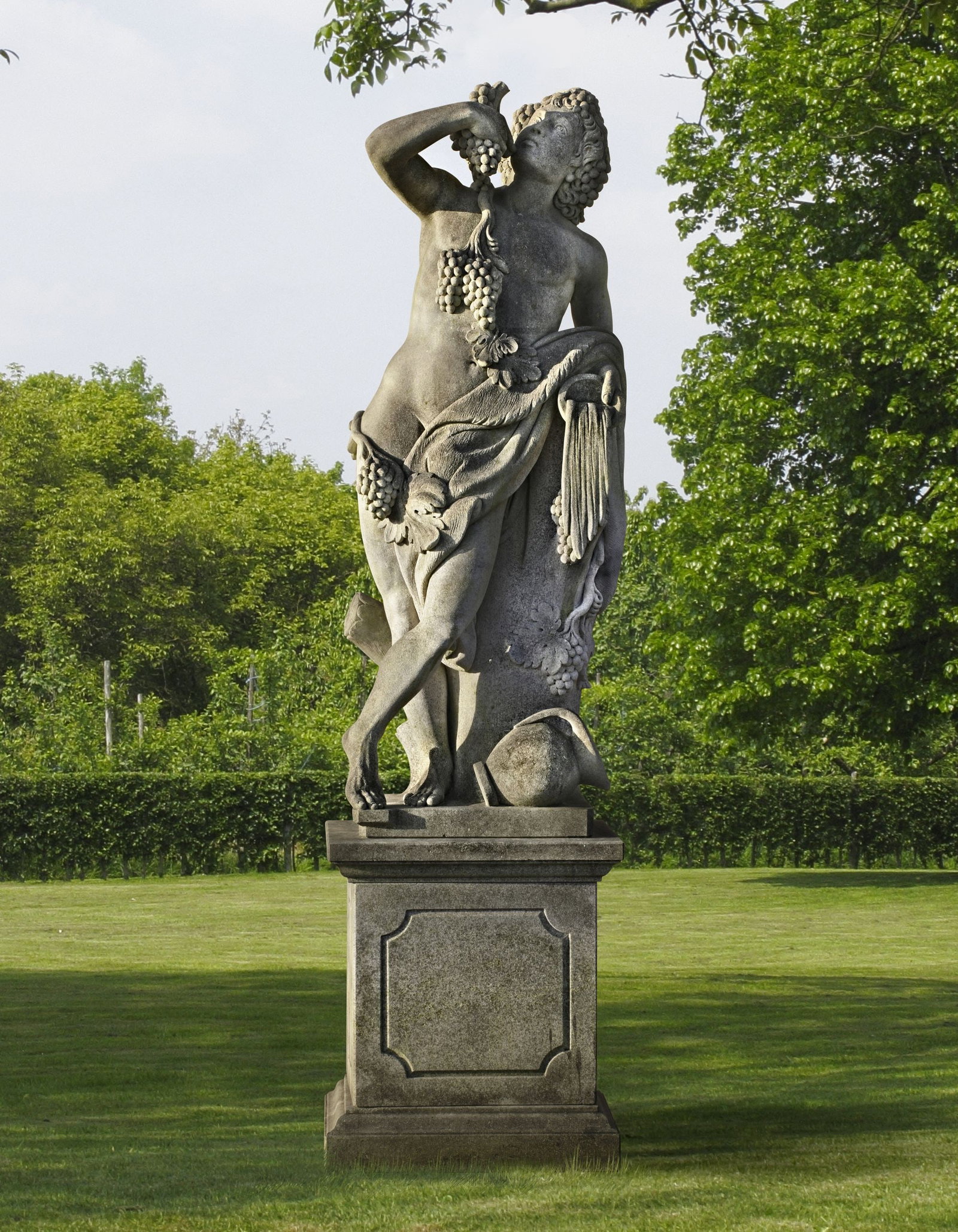 A SCULPTED LIMESTONE MODEL OF BACCHUS, 20TH CENTURY: Bacchus, the Roman god of wine, recognizable by grapes, wine symbols, and a festive appearance. Height overall 260 cm. Height pedestal 80 cm., square 70 by 60 cm.
