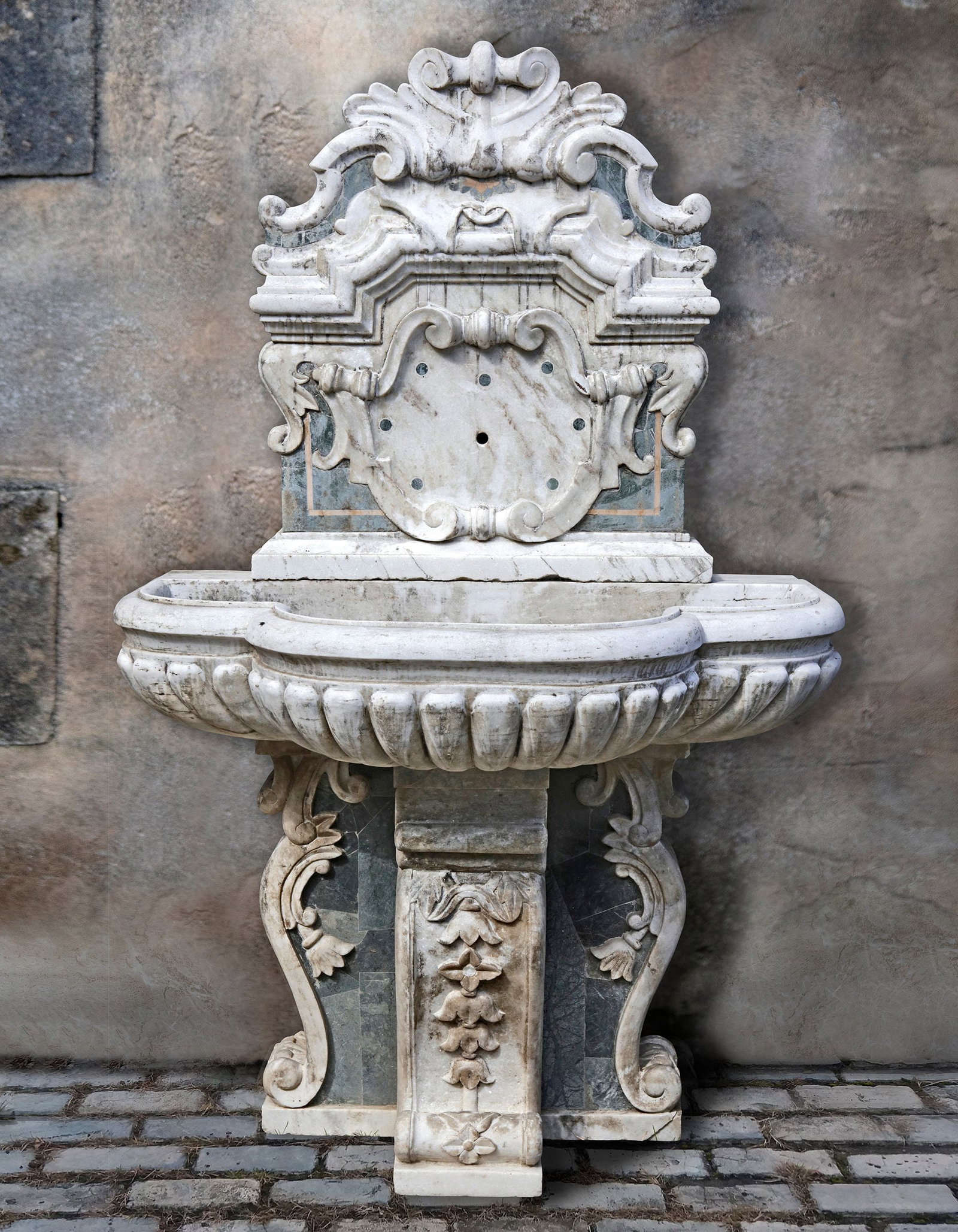 A CARVED WHITE AND SPECIMEN MARBLE WALL FOUNTAIN, 19TH CENTURY: With scrolled upper section and central cartouche containing the spout, above a projecting break front ba- sin, the whole with yellow banded dark blue stone inserts. Height 193 cm. Width 121 cm. Depth