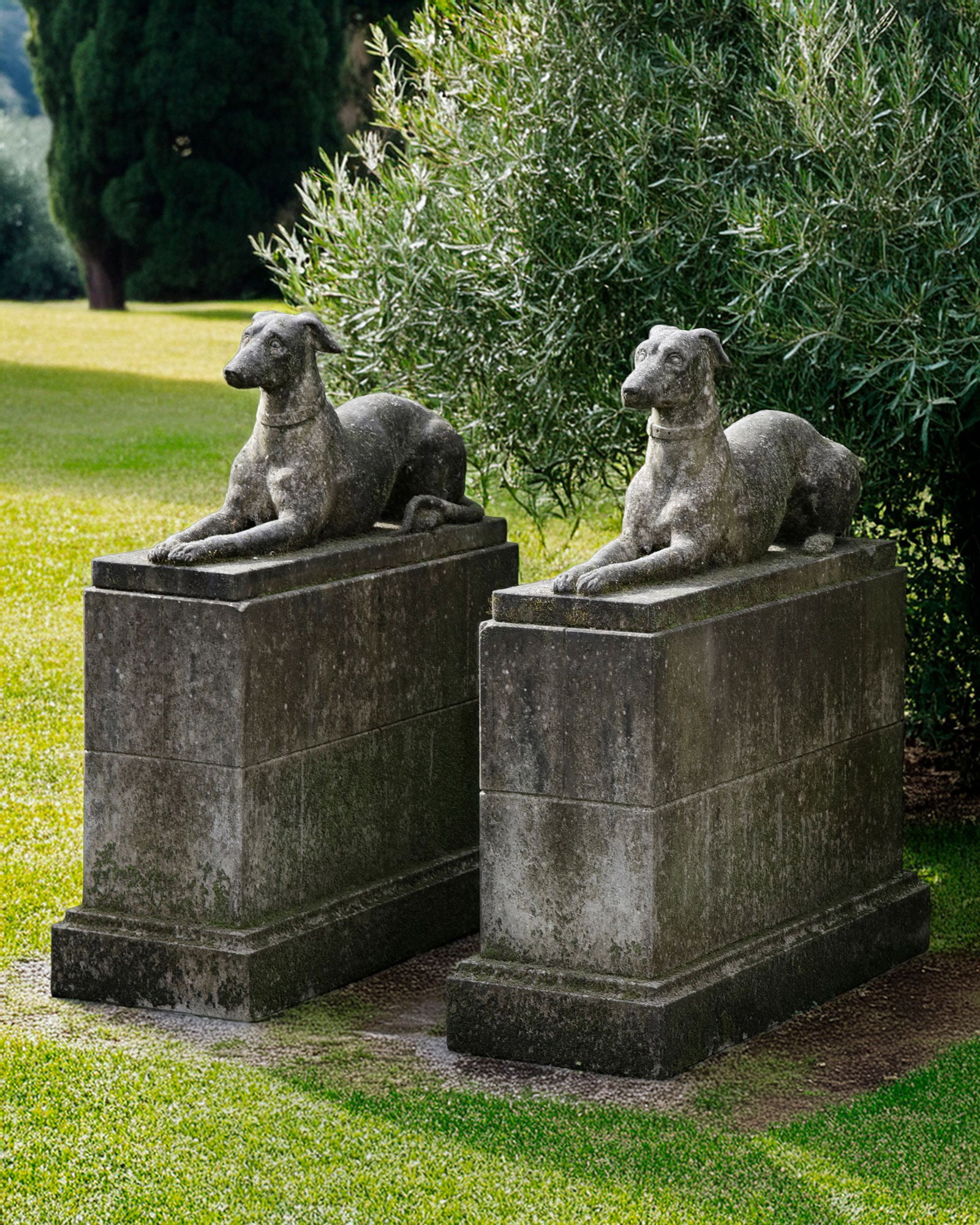 A PAIR OF SCULPTED LIMESTONE MODELS OF RECUMBENT HOUNDS: Each portrayed looking straight forward with forelegs and paws stretched out. Height 47,5 cm. Length 97 cm. Width 29 cm. Set on a rectangular section block work pedestals, height 71,7 cm., length 108 