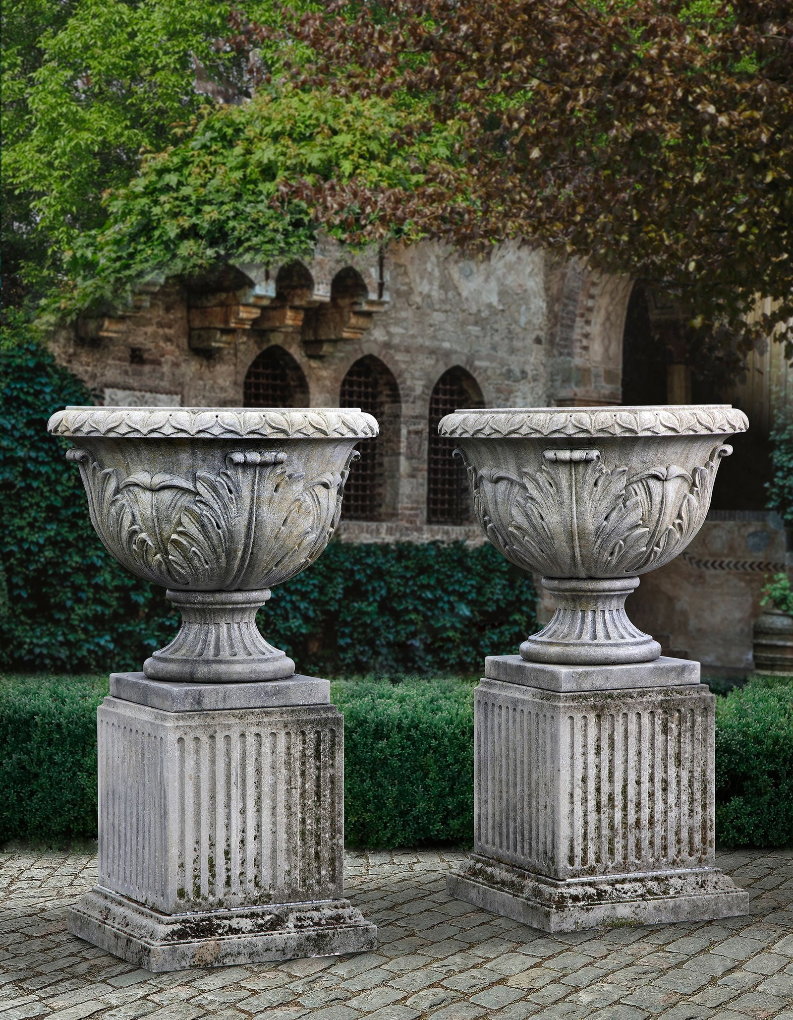 A PAIR OF CARVED LIMESTONE URNS ON PEDESTALS, LATE 20TH CENTURY: With stiff carved decoration with acanthus leaves on the body above fluted pedestals. Height overall 135 cm. Diameter 81 cm. Square at the base 59 cm.