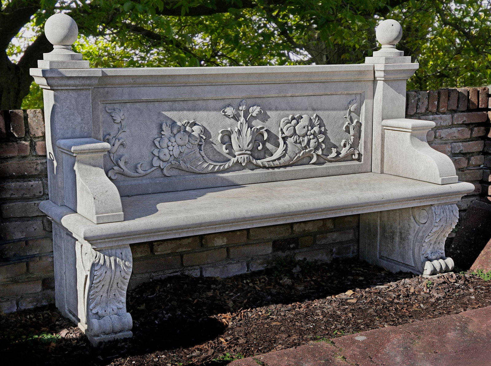 A CARVED LIMESTONE GARDEN BENCH, LATE 20TH CENTURY: This elegant stone garden bench with classic look is carved of limestone. The backrest features the horn of abundance. At either end are square pillars topped with round stone balls. The armrests are 