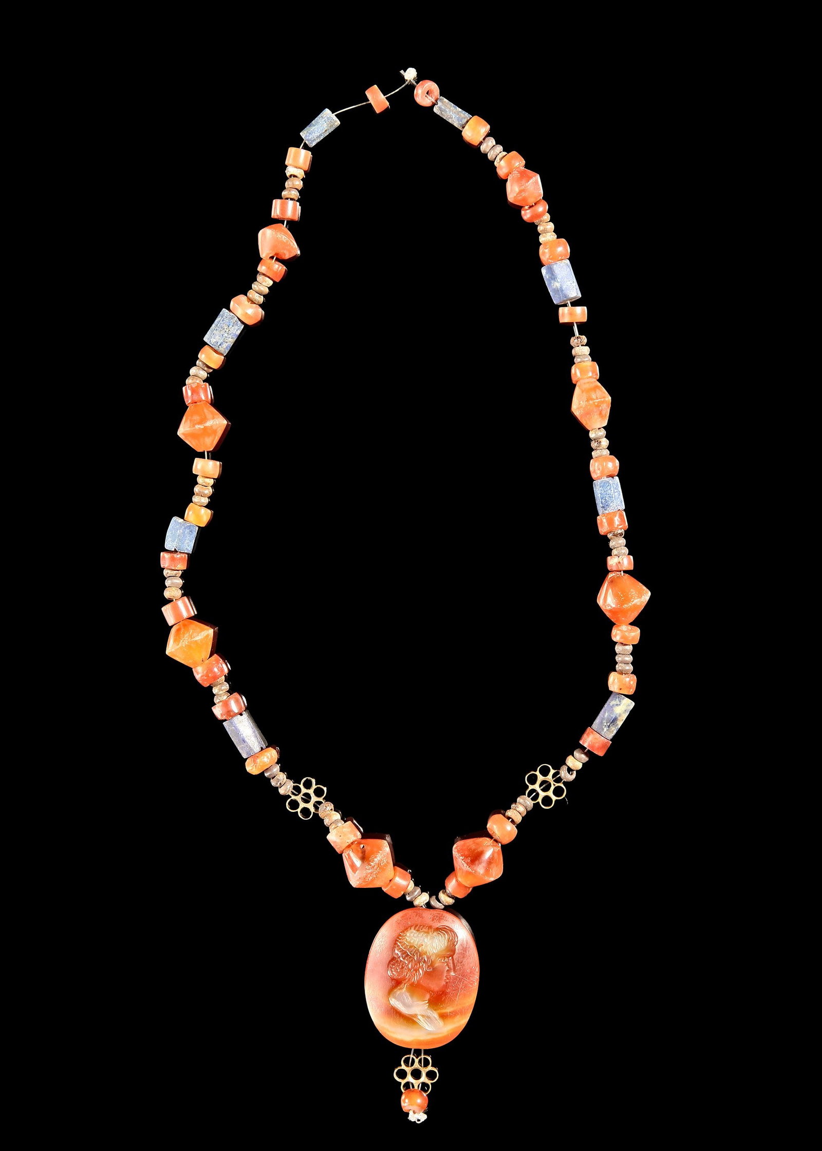 A CARNELIAN CAMEO NECKLACE WITH LAPIS ACCENTS: A striking strand of ancient carnelian in rich amber and orange hues. Faceted, barrel, and disc carnelian beads are paired with deep blue lapis lazuli cylinders and small earthy spacers. Centered by l