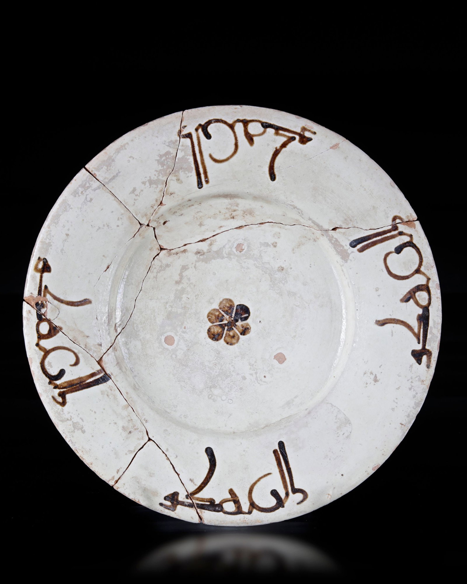 A SLIP-PAINTED POTTERY DISH, PERSIA, 10TH CENTURY: Of shallow form with wide, slightly flaring rim on a stepped foot decorated in manganese on a cream ground with a rosette to the well, the rim with four inscriptions. Diameter 25.6 cm.