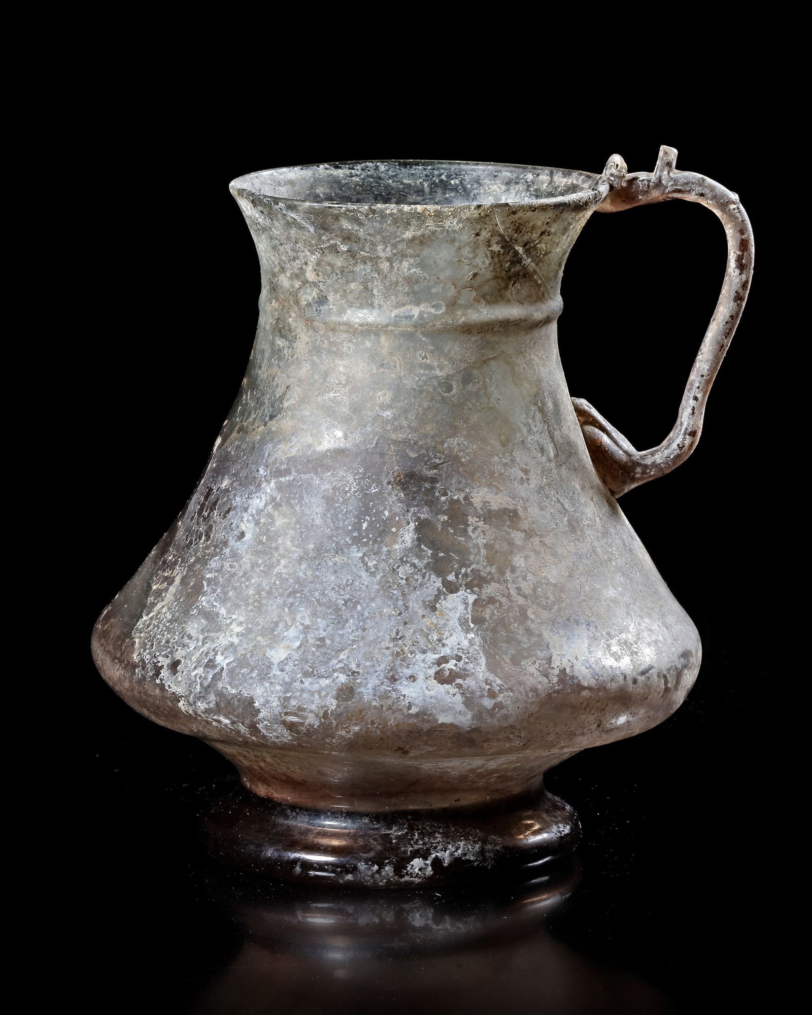 AN IRIDESCENT GLASS JUG, PERSIA, 10TH-11TH CENTURY: Finely blown glass jug of elegant, flaring form, with a narrow neck and gently expanding body, raised on a small ring foot and fitted with a curved handle. the surface displays a striking iridescent s