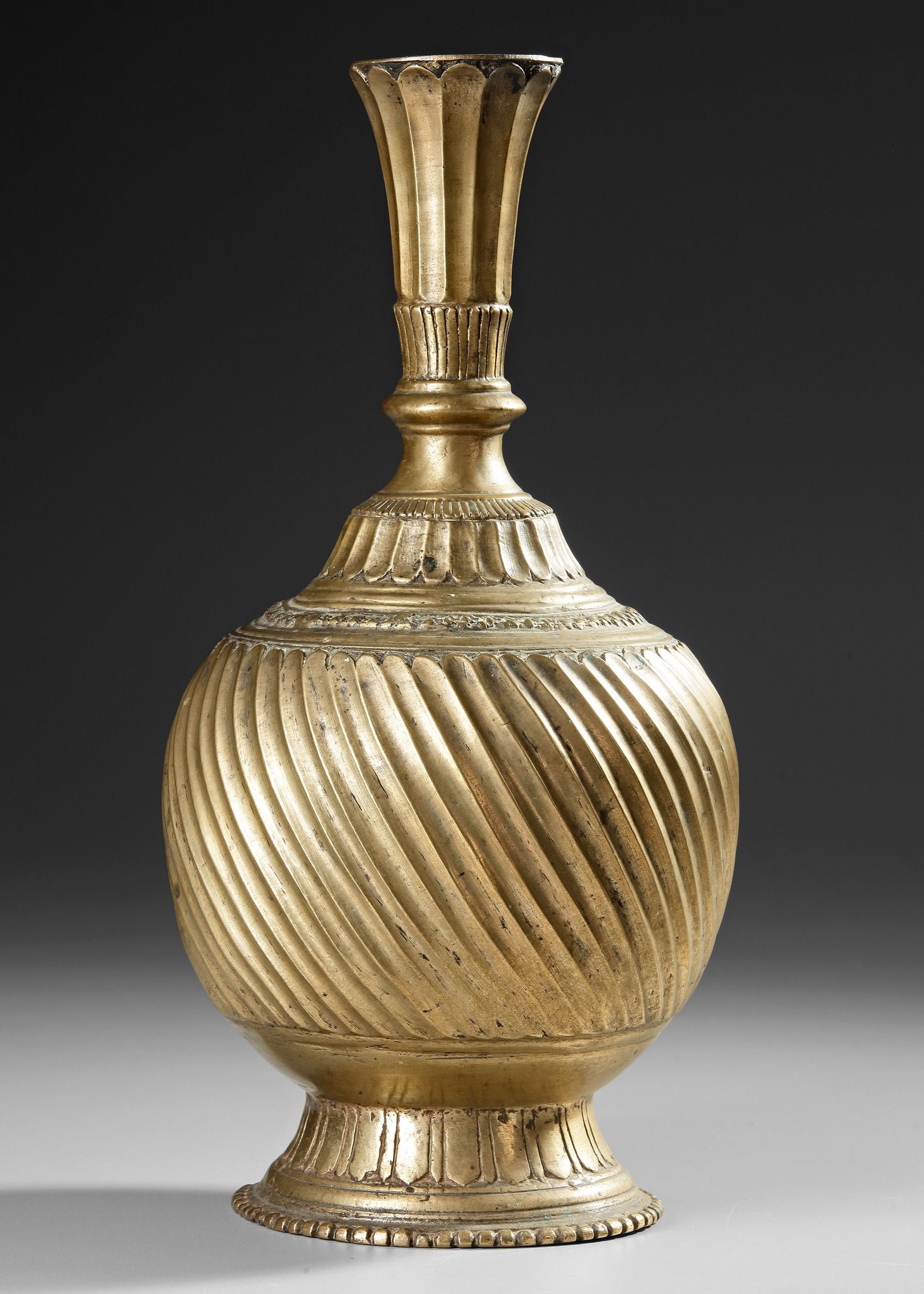 A FINE MUGHAL GILT-BRONZE VASE, INDIA, 17TH CENTURY: Of elegant baluster form, the globular body rising from a spreading, lotus-petal foot to a waisted neck terminating in a flared, petal-lobed mouth. The surface is richly ornamented with bold spiral ga
