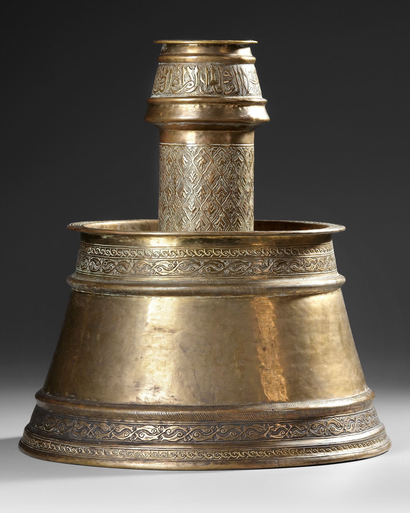 A FINE OTTOMAN GILT-BRASS CANDLESTICK, LATE 18THâ€“EARLY 19TH CENTURY: A monumental gilt-brass mosque candlestick of architectural form, with a broad circular drip pan above a truncated conical base supporting a tall cylindrical candle socket. The socket is engraved with