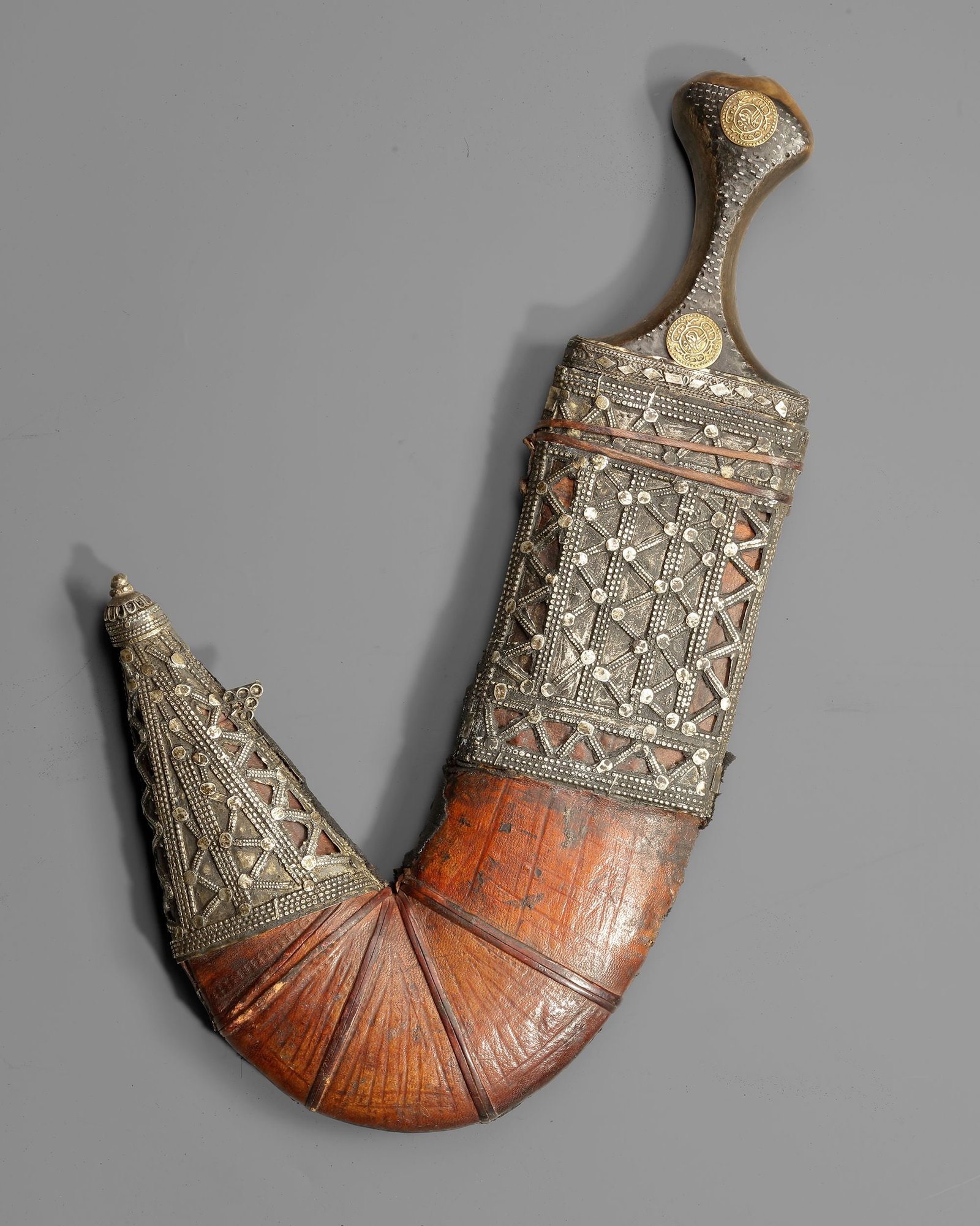 A SILVER YEMENI DAGGER, EARLY 20TH CENTURY: A traditional Yemeni jambiya featuring a sharply curved steel blade and a finely decorated silver hilt set with gold medallion ornaments. The scabbard is covered in leather and embellished with elabor