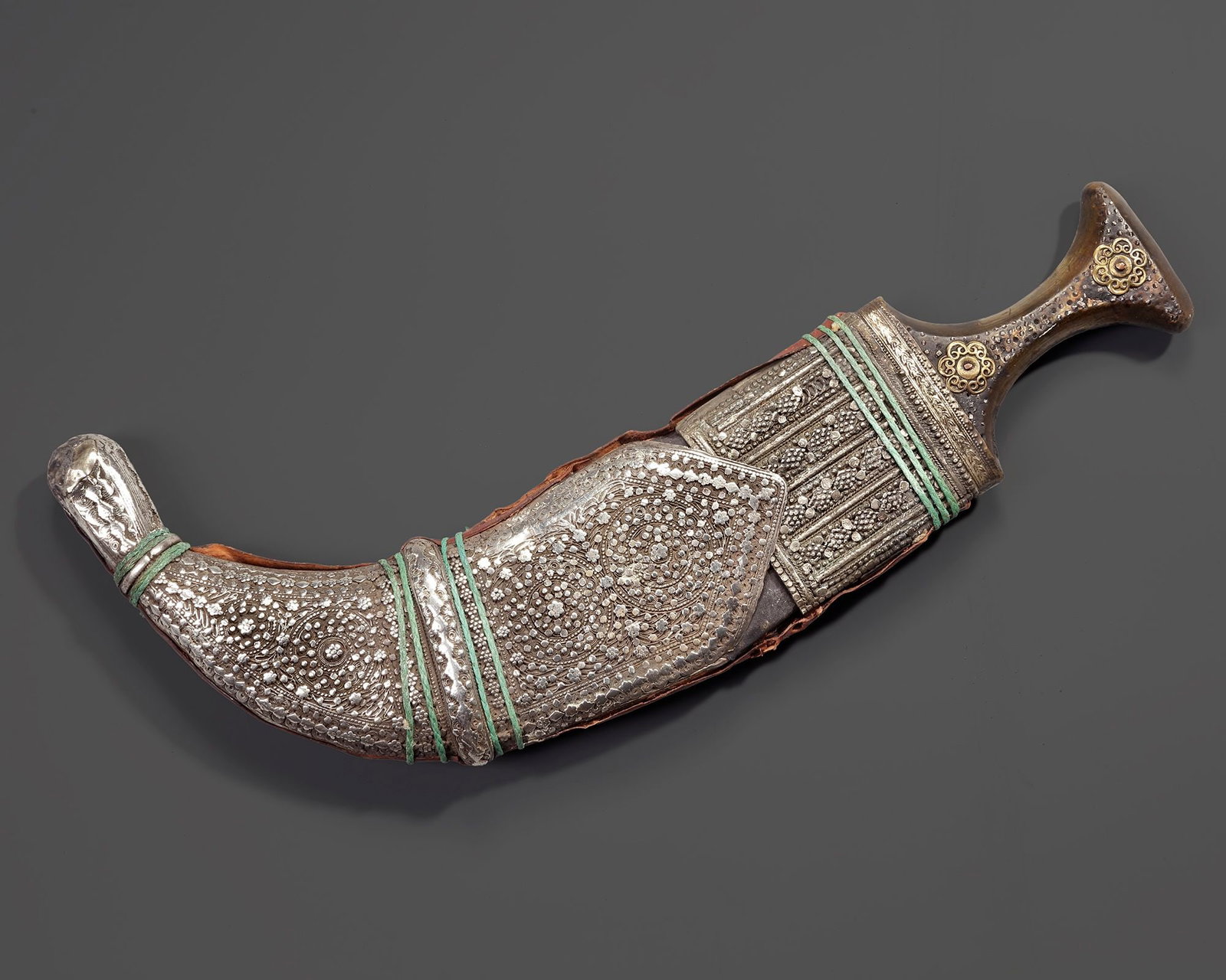 A SILVER YEMENI DAGGER (JANBIYA,) EARLY 20TH CENTURY: A traditional Yemeni janbiya with a pronounced curved form, featuring a finely worked silver scabbard decorated with intricate floral and geometric motifs. The dagger is mounted with a wooden hilt and