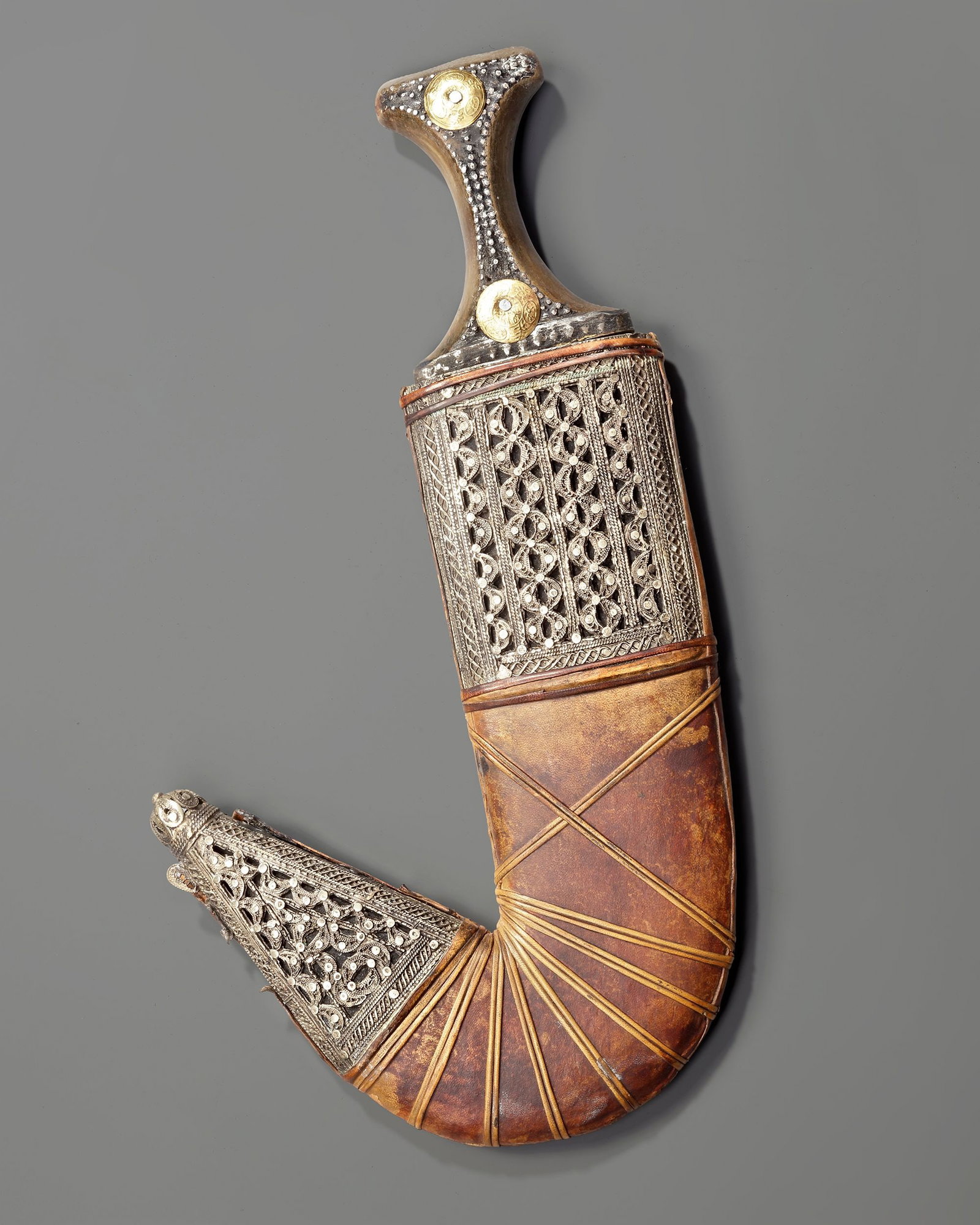 A SILVER YEMENI DAGGER, EARLY 20TH CENTURY: A traditional Yemeni jambiya featuring a sharply curved steel blade and a finely decorated silver hilt set with gold medallion ornaments. The scabbard is covered in leather and embellished with elabor