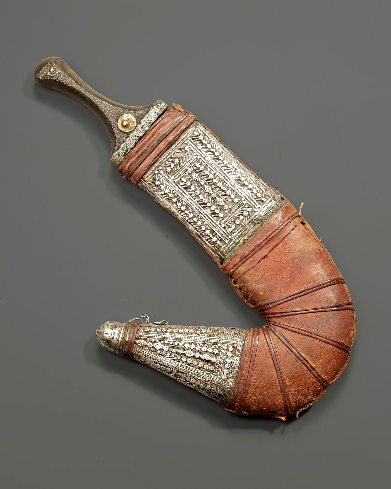 A SILVER YEMENI DAGGER, EARLY 20TH CENTURY: A traditional Yemeni jambiya featuring a sharply curved steel blade and a finely decorated silver hilt set with gold medallion ornaments. The scabbard is covered in leather and embellished with elabor