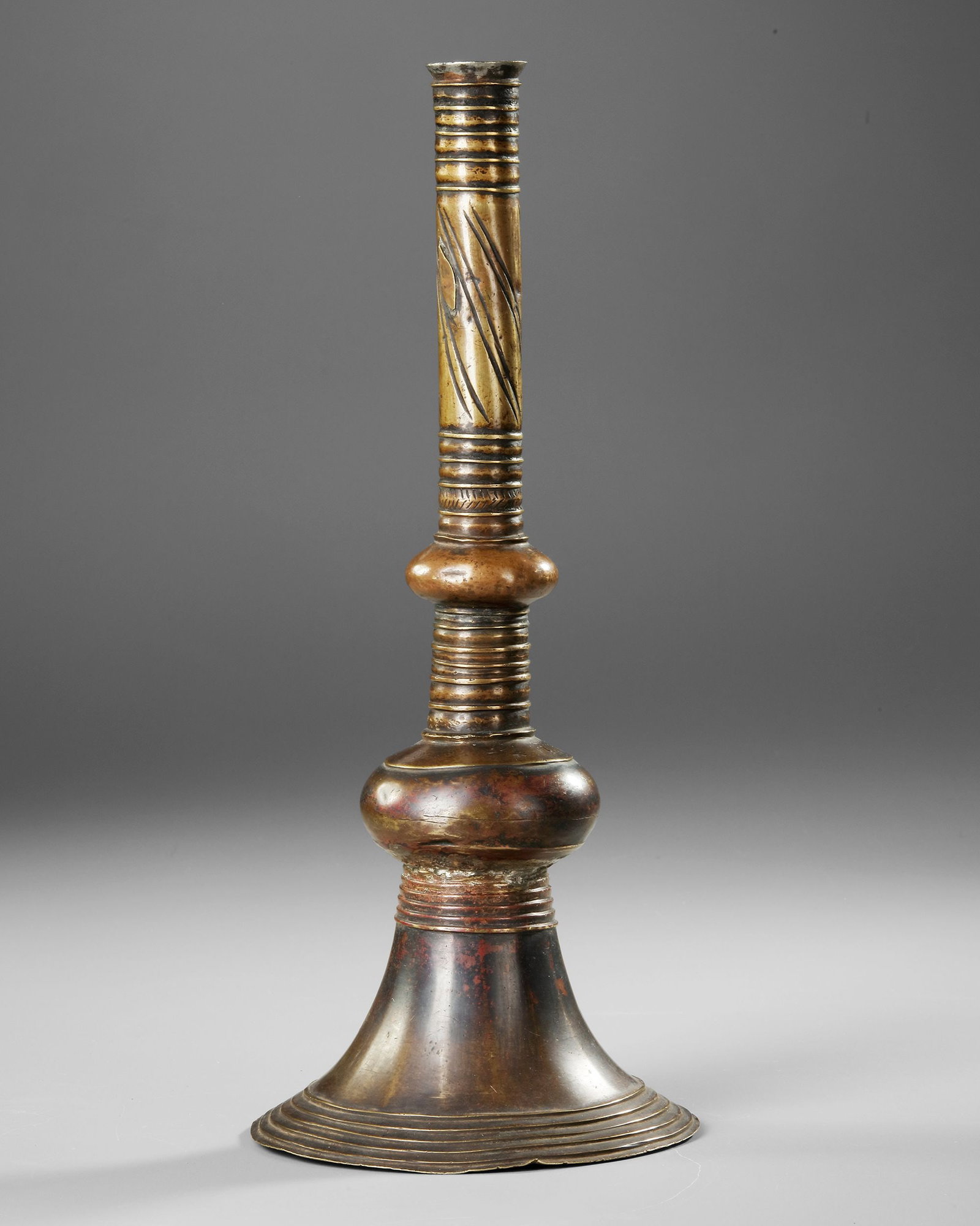 A CANDLESTICK, YEMEN, 16TH CENTURY: Brass candlestick featuring a wide stepped base and a beautifully balanced baluster shaft. The surface displays engraved linear motifs and a rich, naturally aged patina. Height 38 cm. 798 gr.