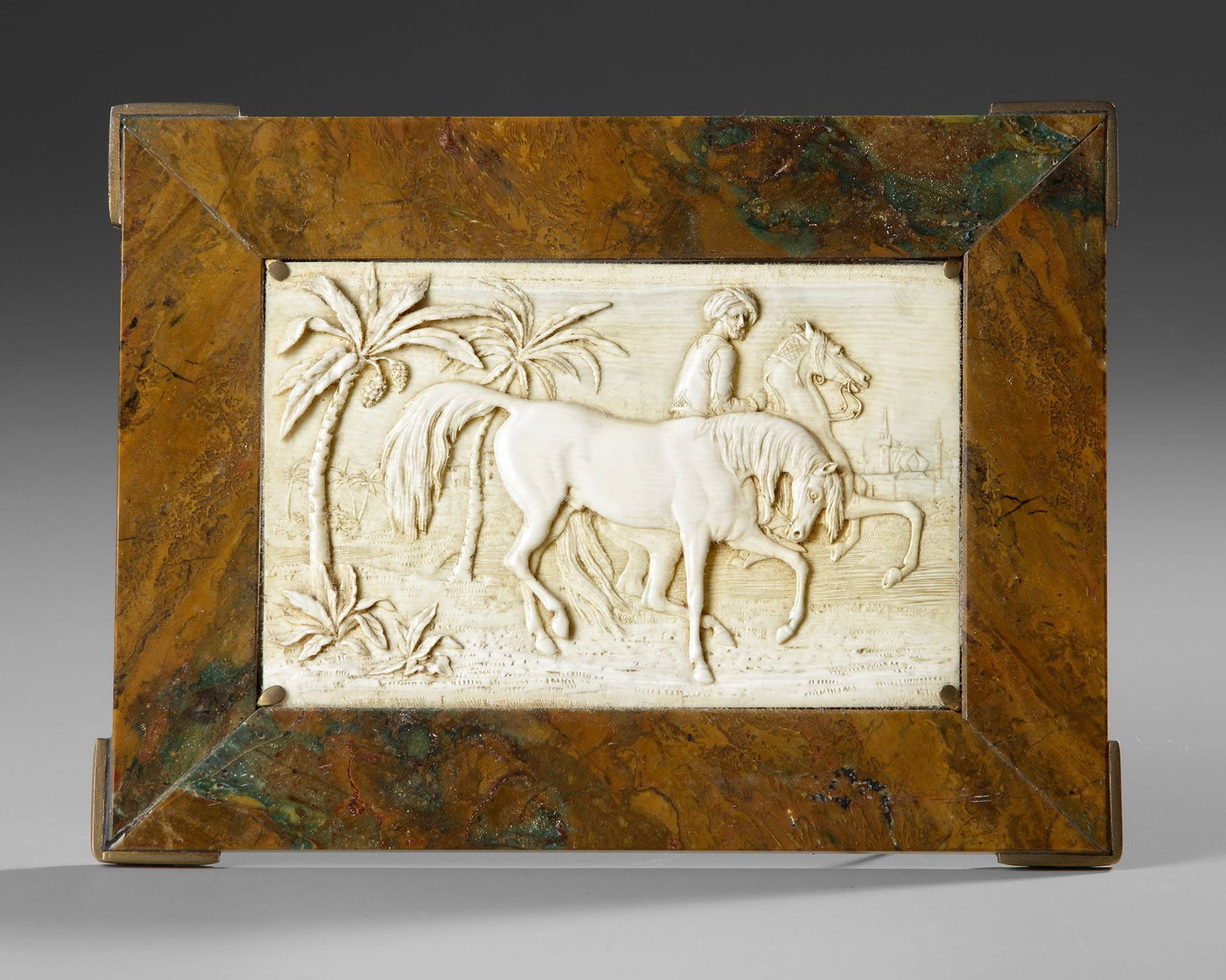 A CARVED BONE RELIEF PANEL, 20TH CENTURY: A finely carved bone relief panel mounted within a brass frame, depicting a horseman and a spirited horse in a palm-filled landscape. Rich in detail and craftsmanship, the scene evokes the romantic im