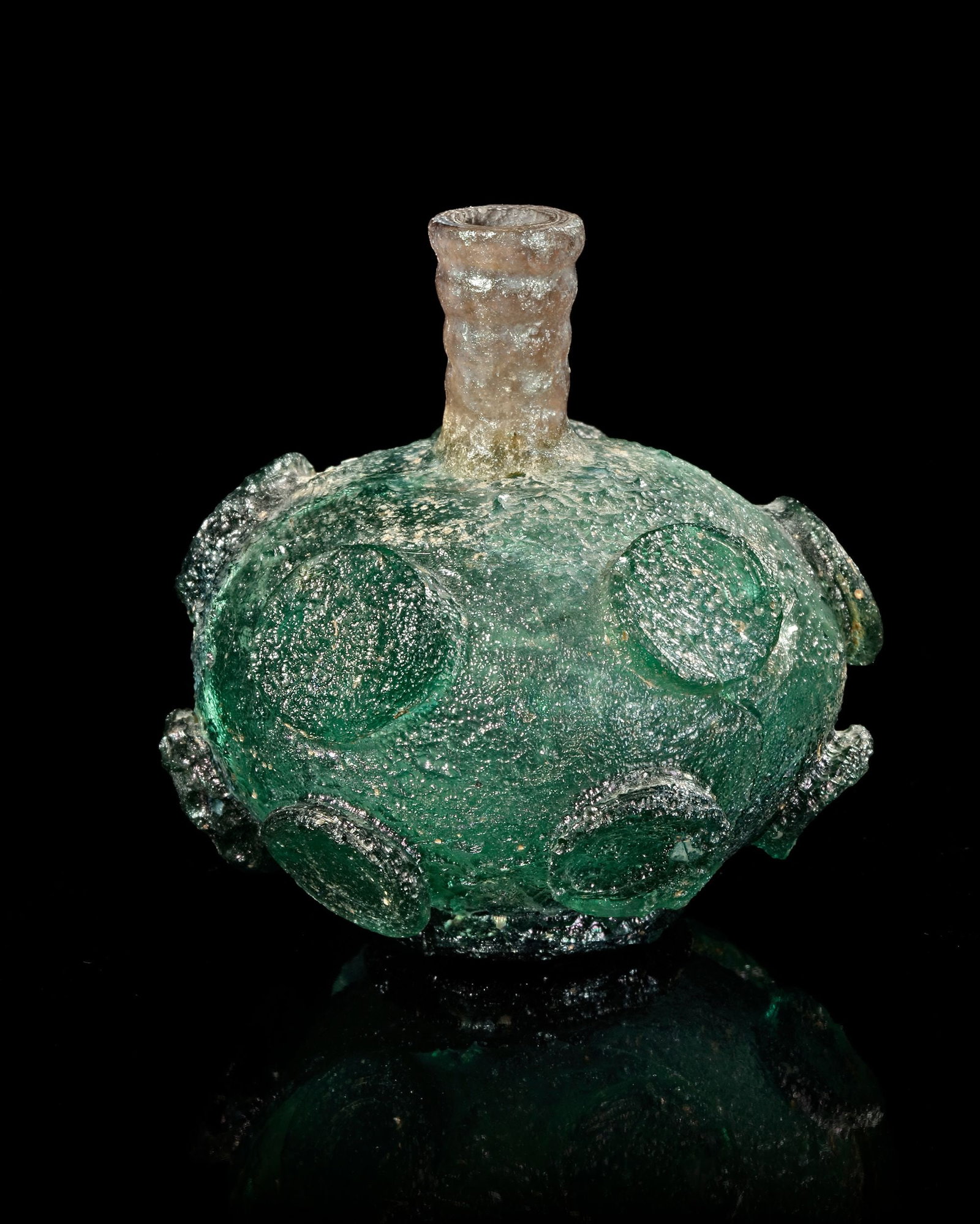 A GLASS FLASK, PERSIA, 10THâ€“11TH CENTURY: A rare and finely crafted medieval Persian glass flask featuring a globular body adorned with applied circular medallions. The vessel displays a striking green hue with shimmering iridescence, a resul